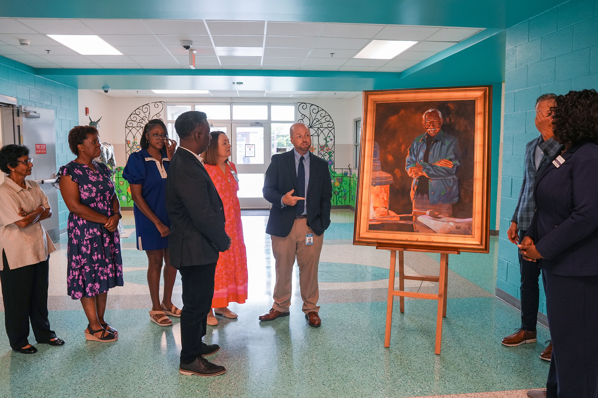 People stand in a circle around a framed portrait on an easel in a hallway with light blue walls.