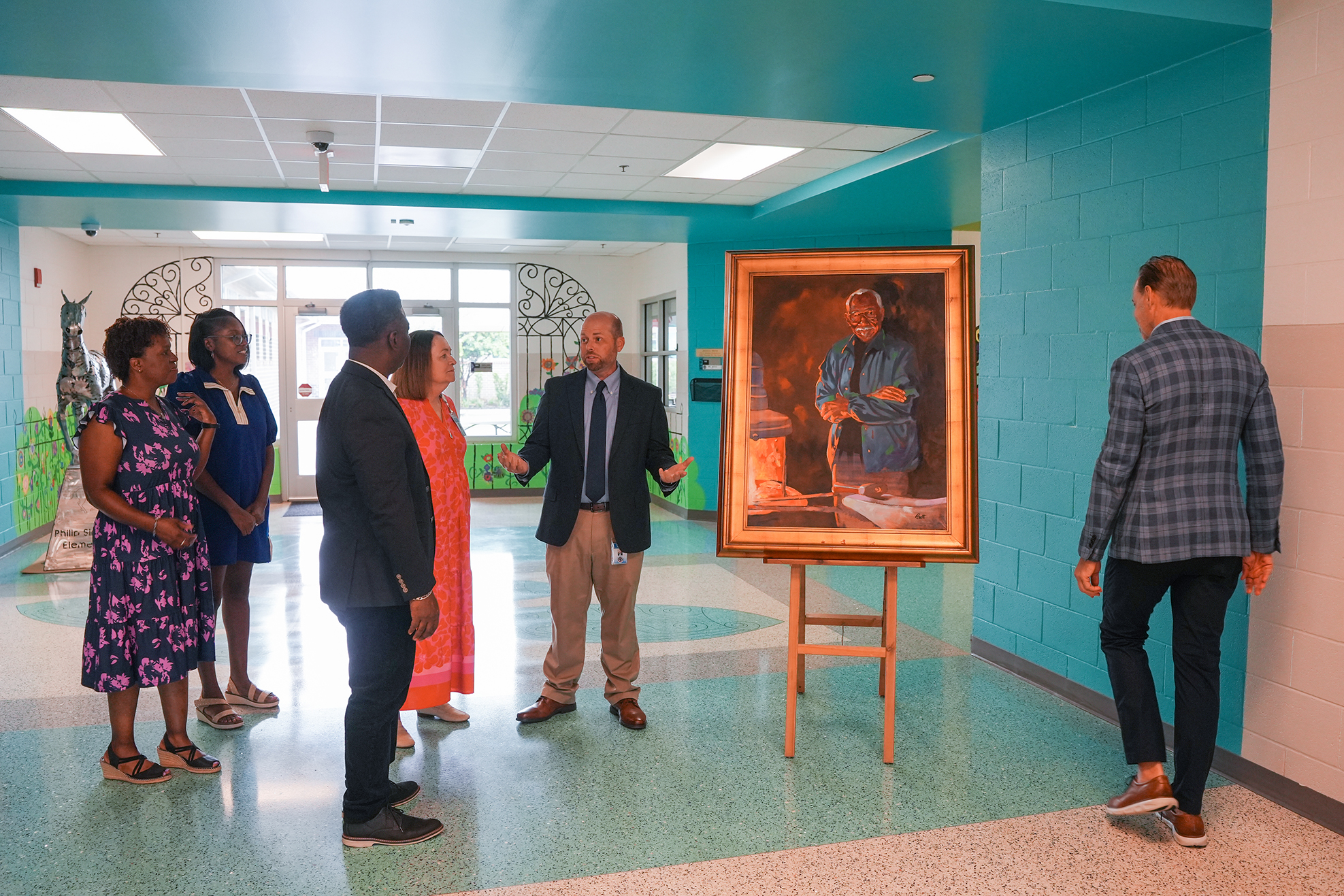 A group of people stand around a framed painting, with a man talking. They are in a hallway with blue walls.