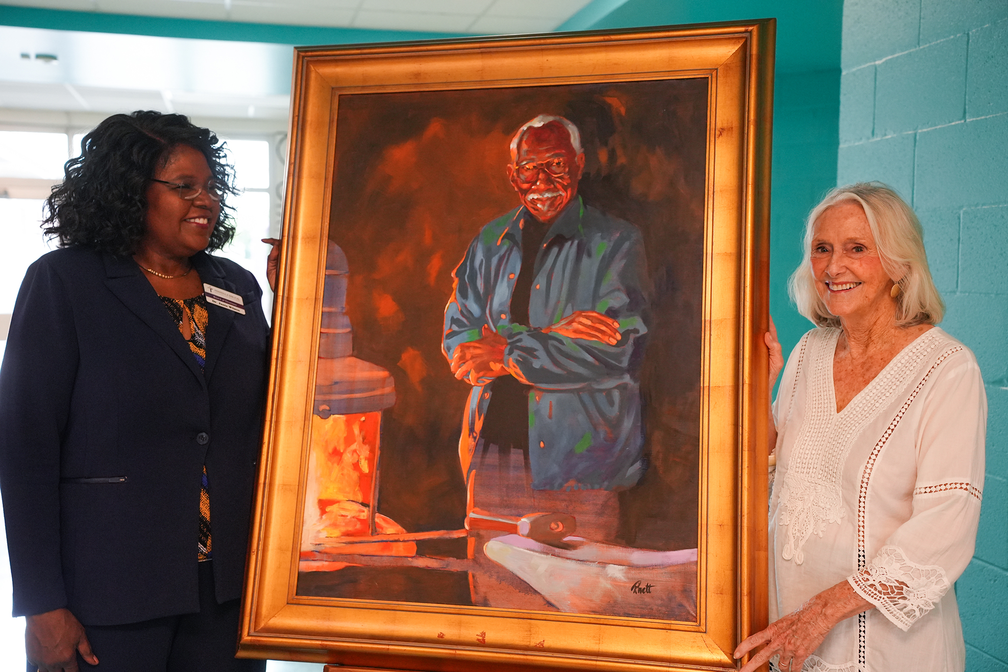 Two women display a framed portrait of a man standing by a fire. One woman wears a business suit, the other a white blouse.