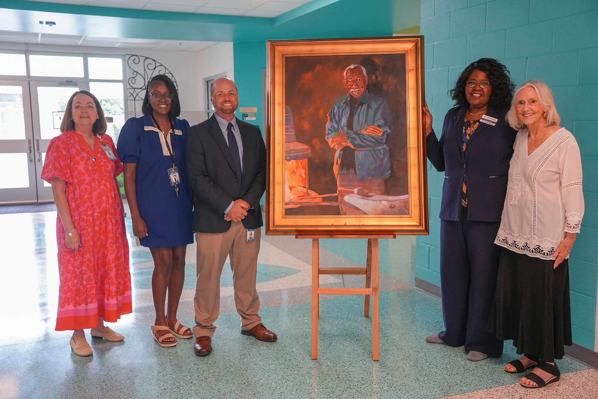 Five people stand in a hallway, two women in dresses and three men in suits, posing with a framed painting.