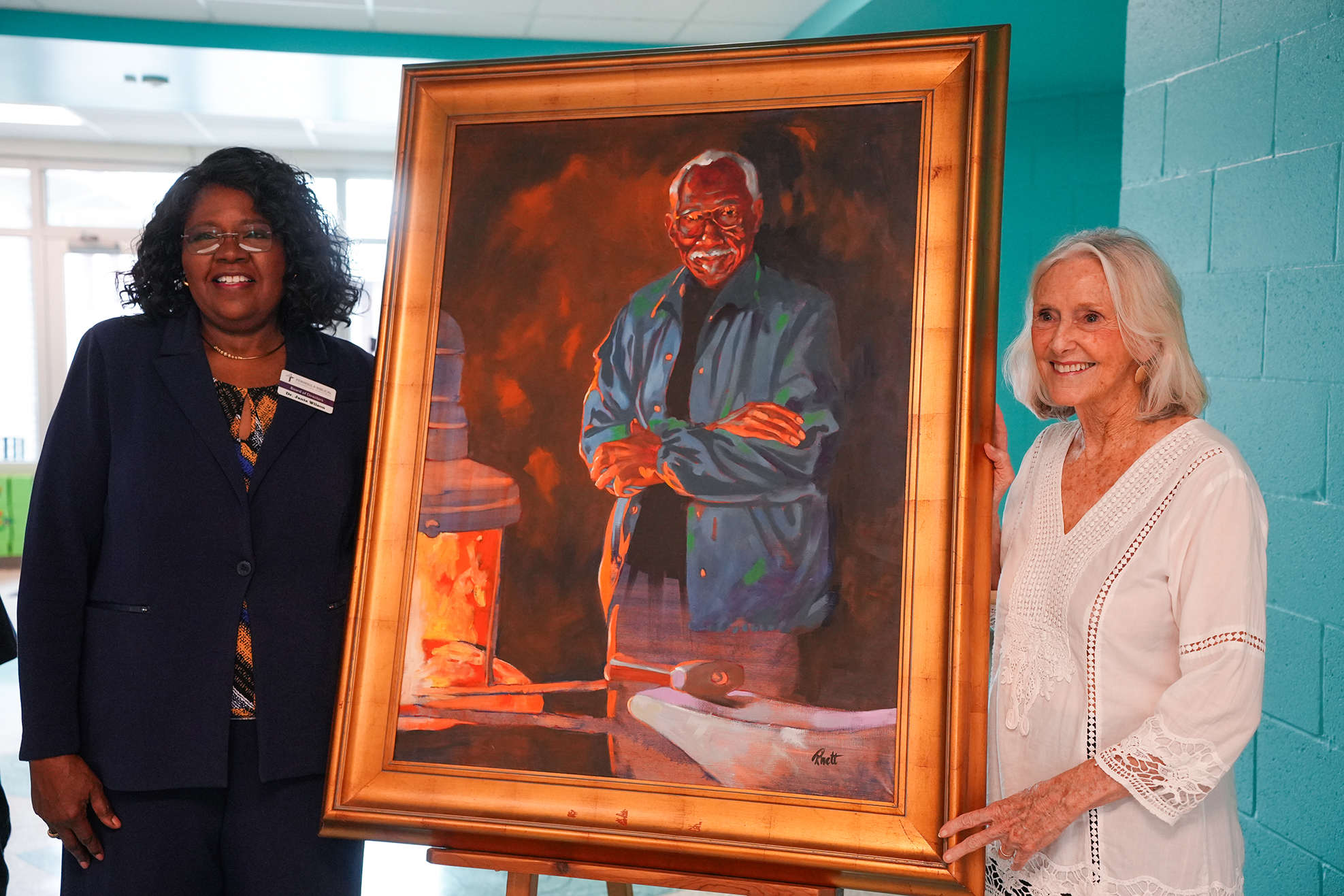 Two women standing next to a large framed portrait of a man with a beard and apron.
