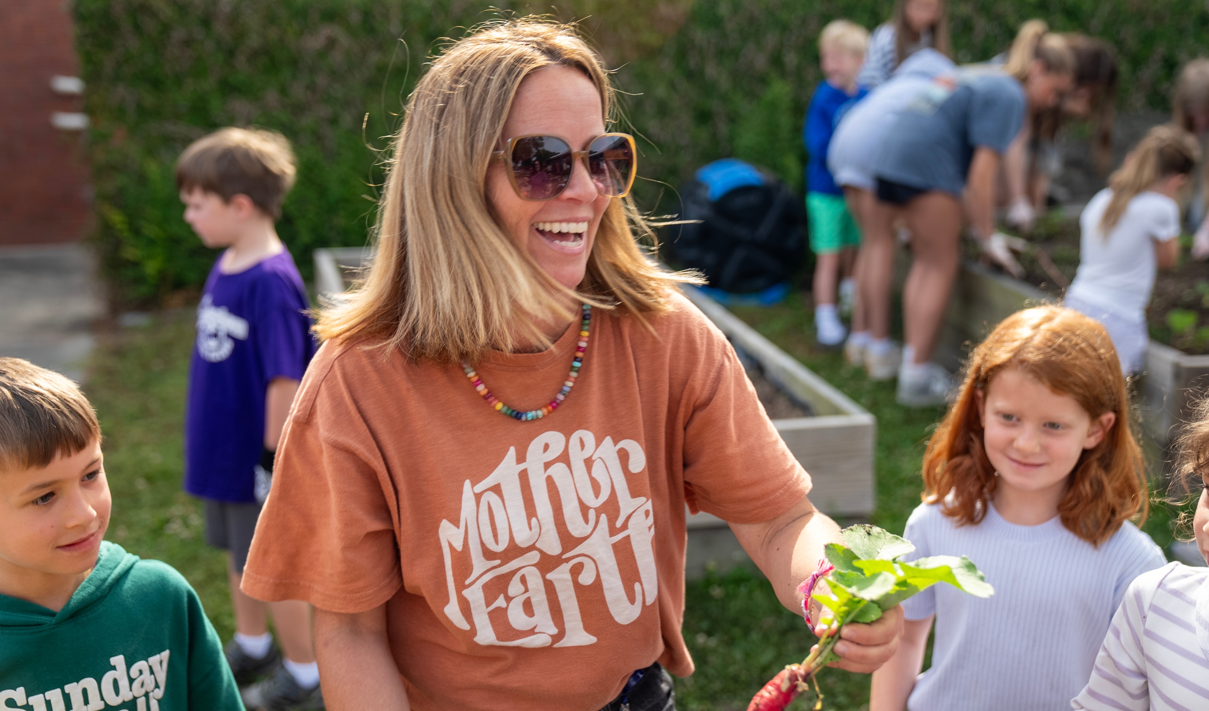 A woman in sunglasses holds a radish, smiling. Behind her, children and adults tend to a garden.