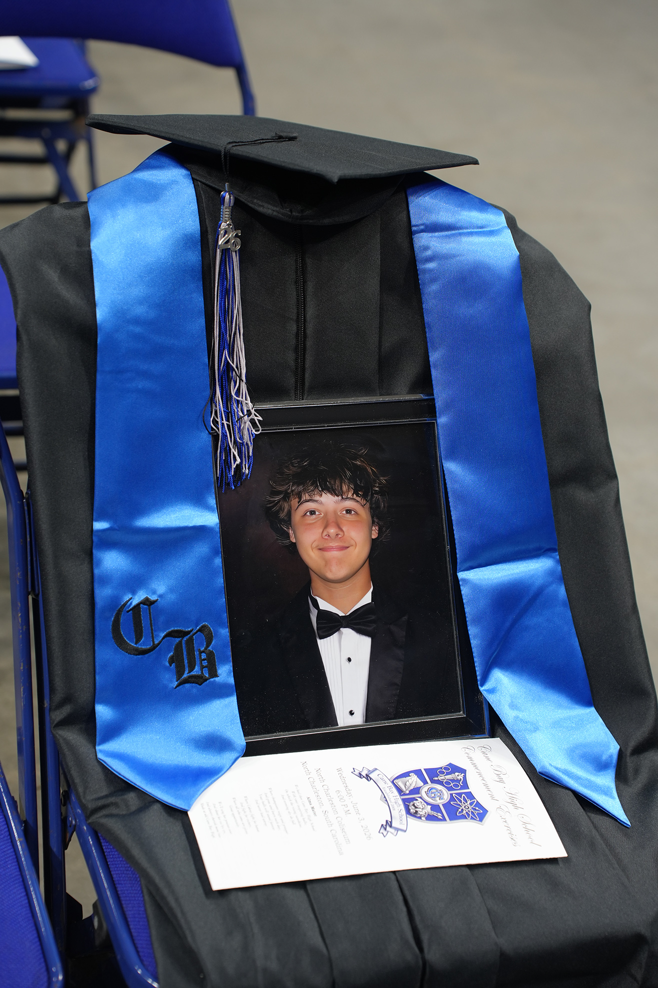 A graduation cap, tassel, and stole sit on a chair. A photo of a young man in a tuxedo and bow tie is framed. A certificate rests on top.