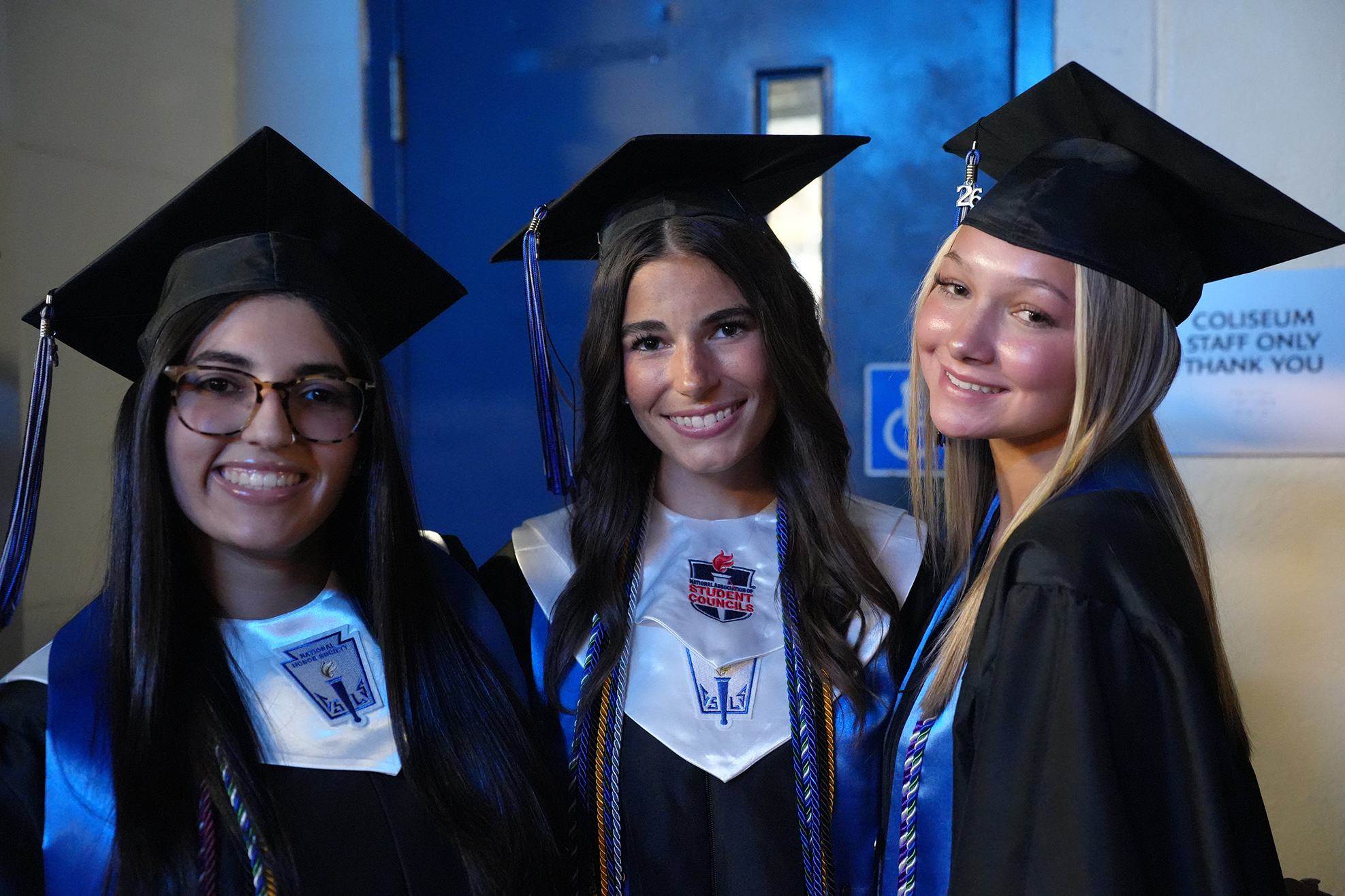 Three women in black graduation gowns and caps, smiling, standing together. Two wear glasses, one has a sash.