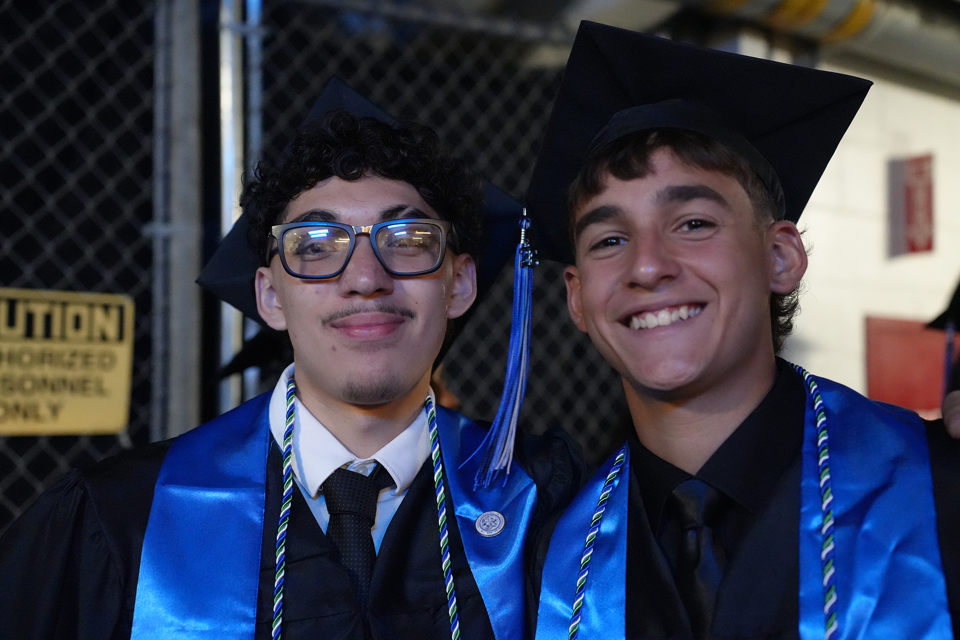 Two young men, both wearing blue graduation gowns, are smiling. One wears glasses, the other a tie.