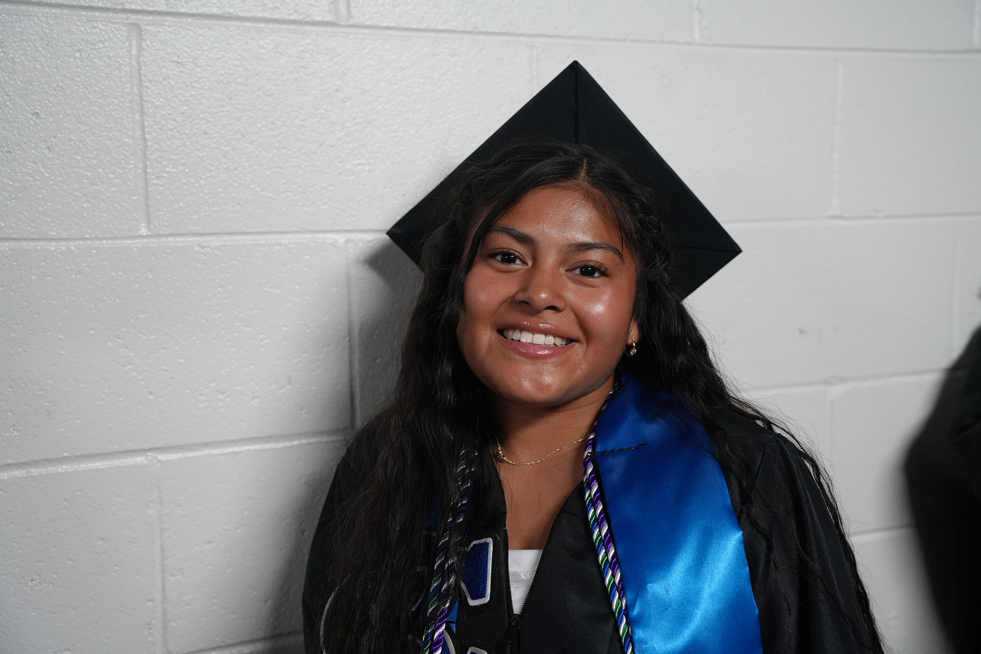 Woman in a cap and gown smiles and stands against a white brick wall.