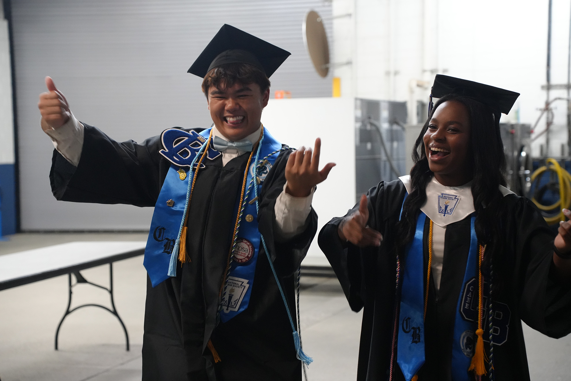 Two people in graduation gowns and caps, one giving a thumbs-up, stand indoors near a white table.