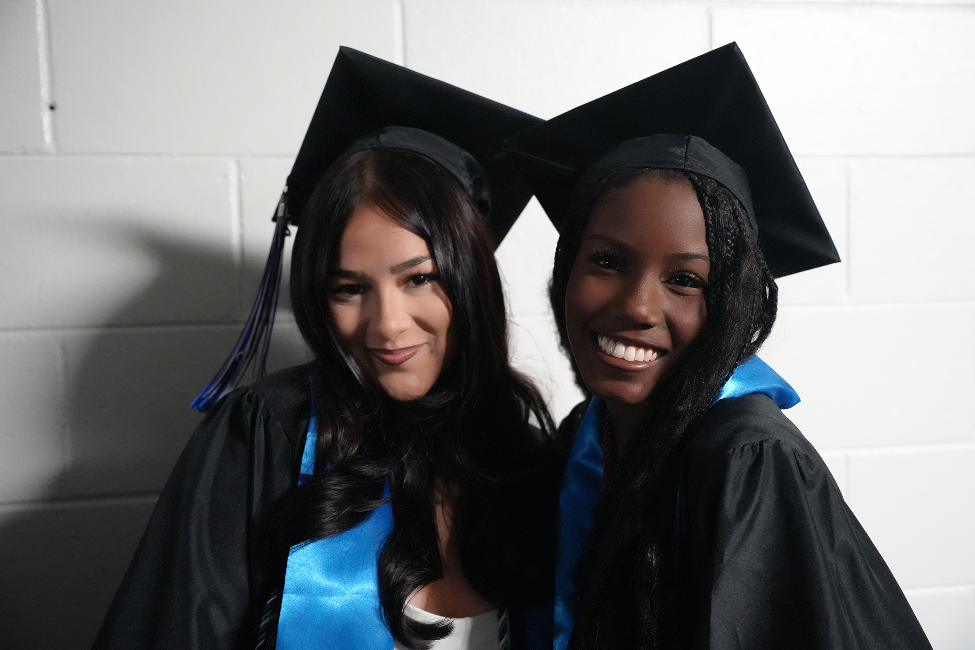 Two women in graduation gowns and caps, one wearing blue trim, smile for a photo against a white brick wall.