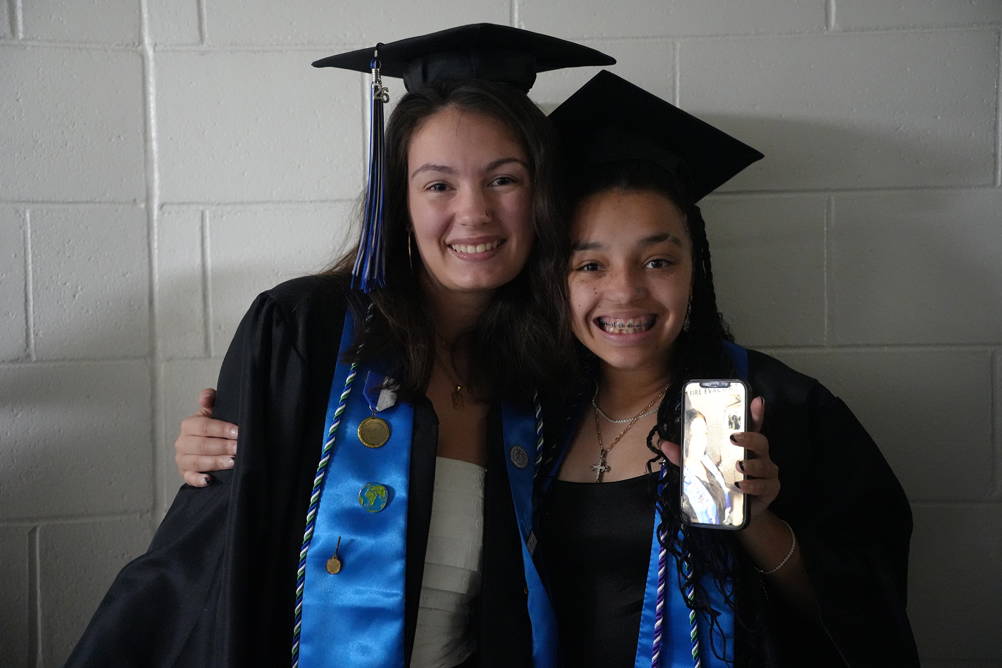 Two women in graduation attire, one holding a phone, posing against a white brick wall.