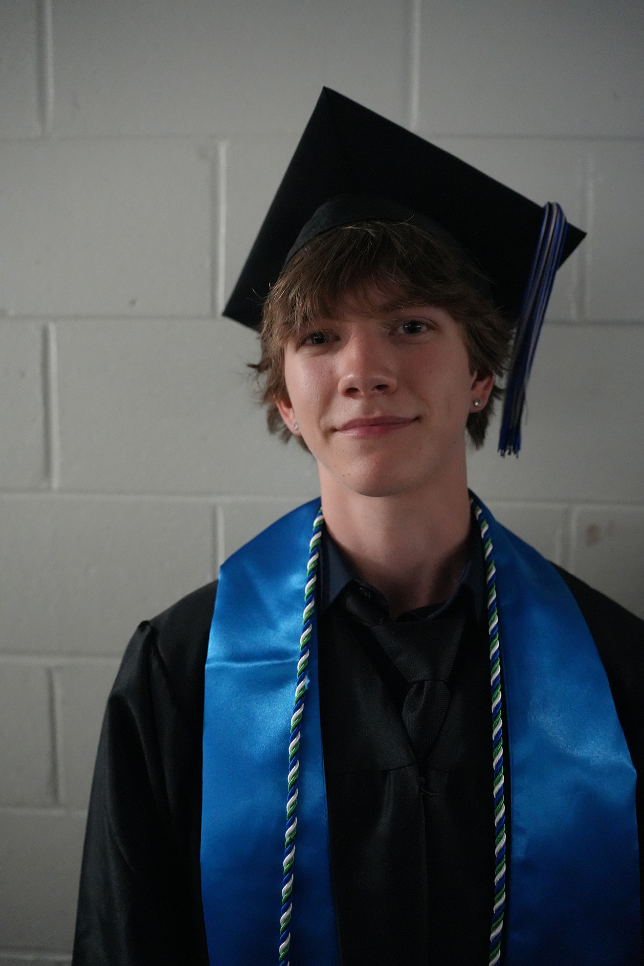 Man in cap and gown, smiling, with a blue graduation stole, standing before a white brick wall.