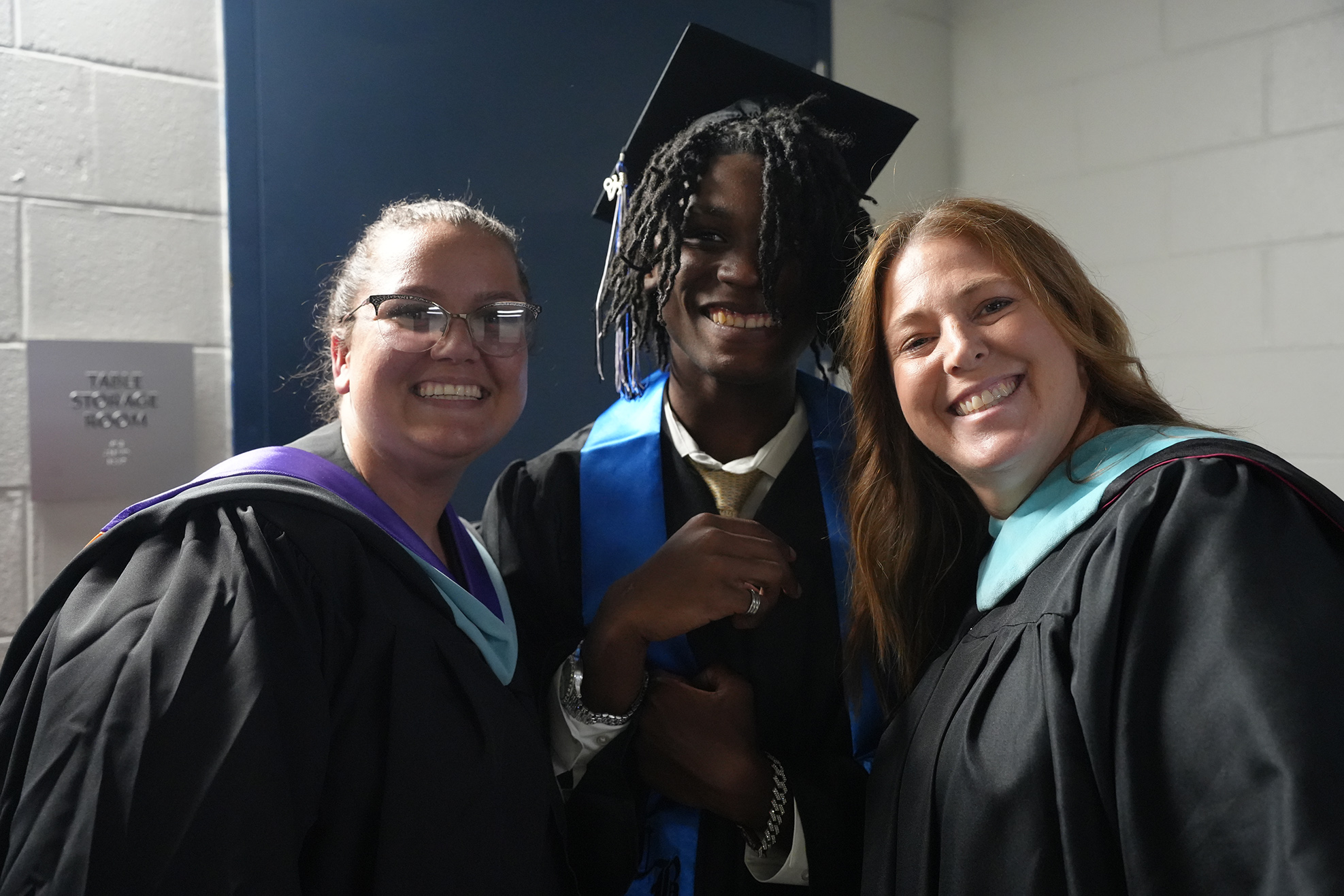 Three individuals in academic regalia stand close together, smiling. One has a graduation cap, another wears glasses, and all wear black gowns.