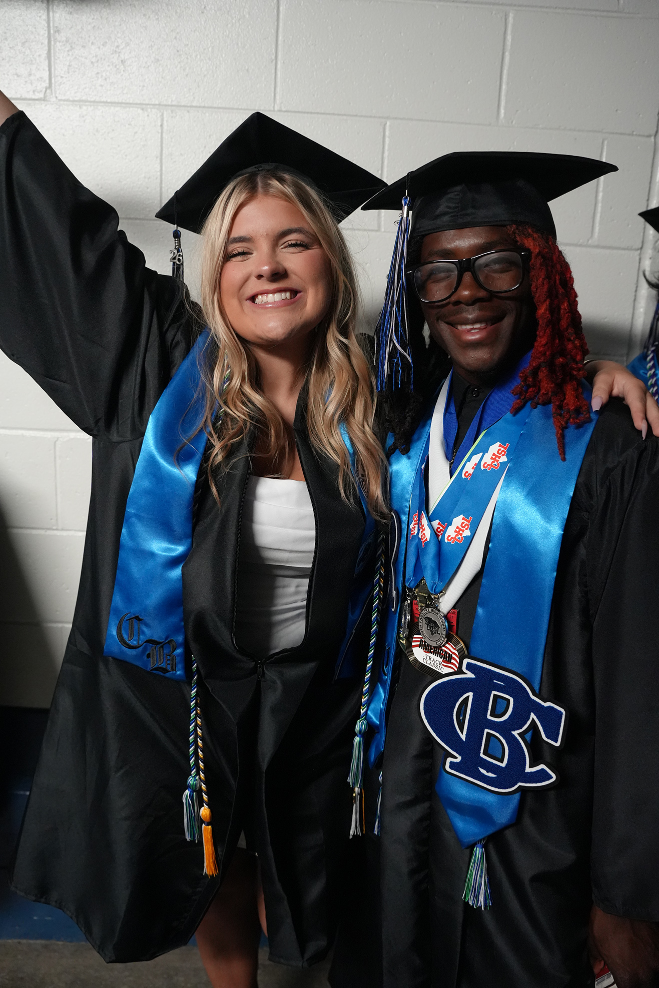Two graduates in black gowns with blue sashes, one with a white inner shirt and the other with a red braid, posing for a photo.