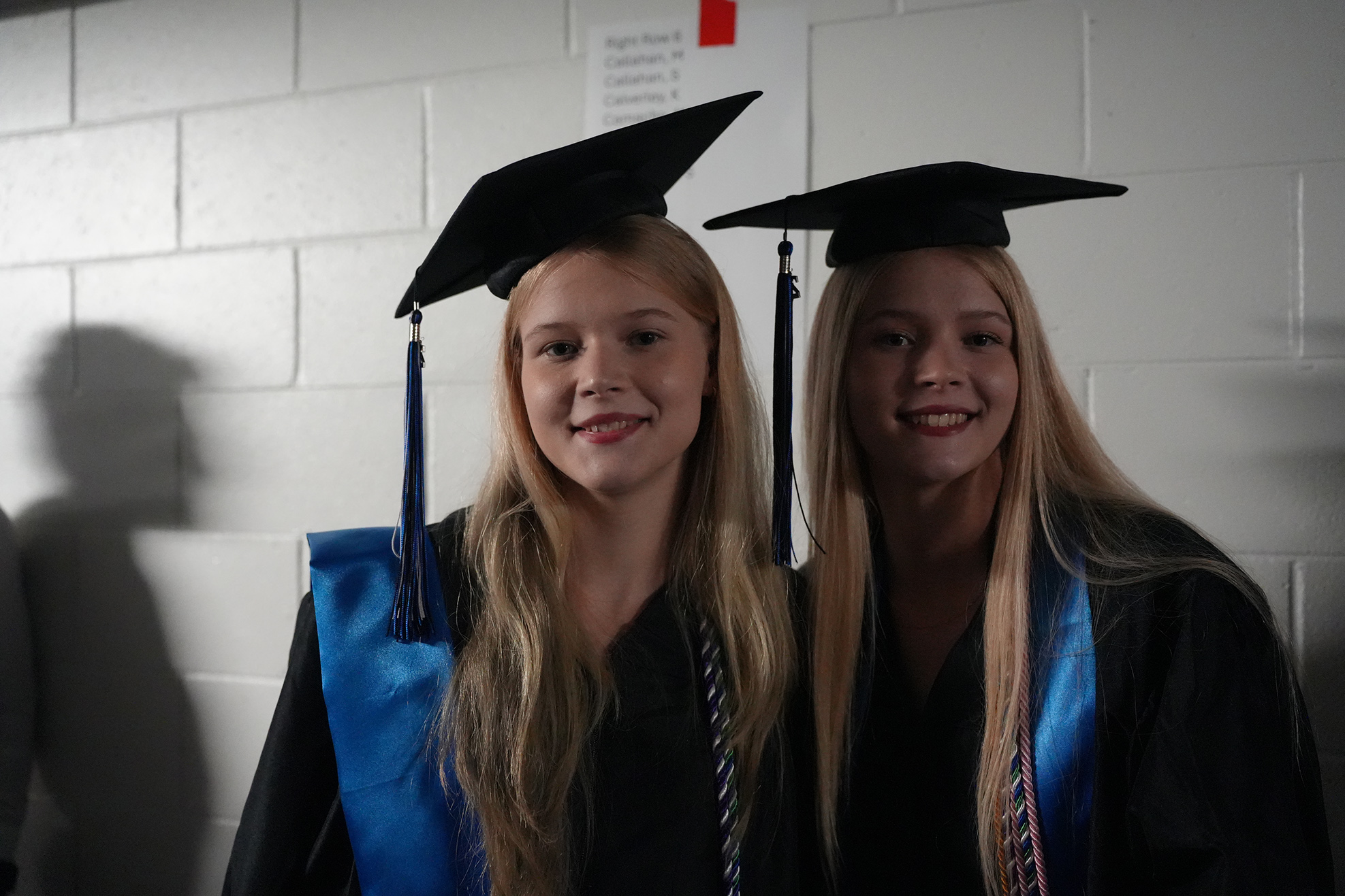 Two women in black gowns and mortarboards stand close, smiling at a white-bricked wall.