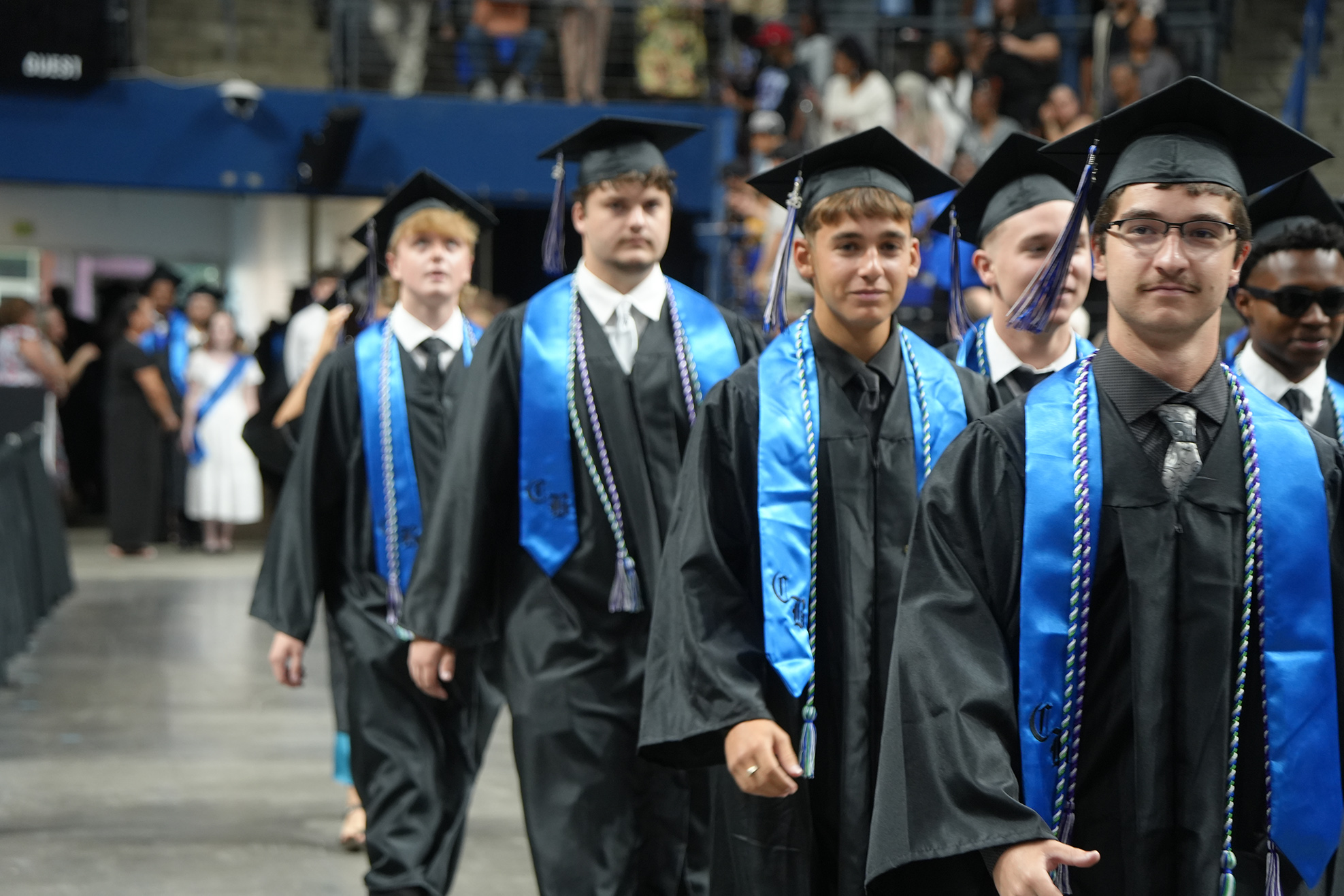 Graduates in black gowns and blue stoles walk in a line on a stage with a crowd.