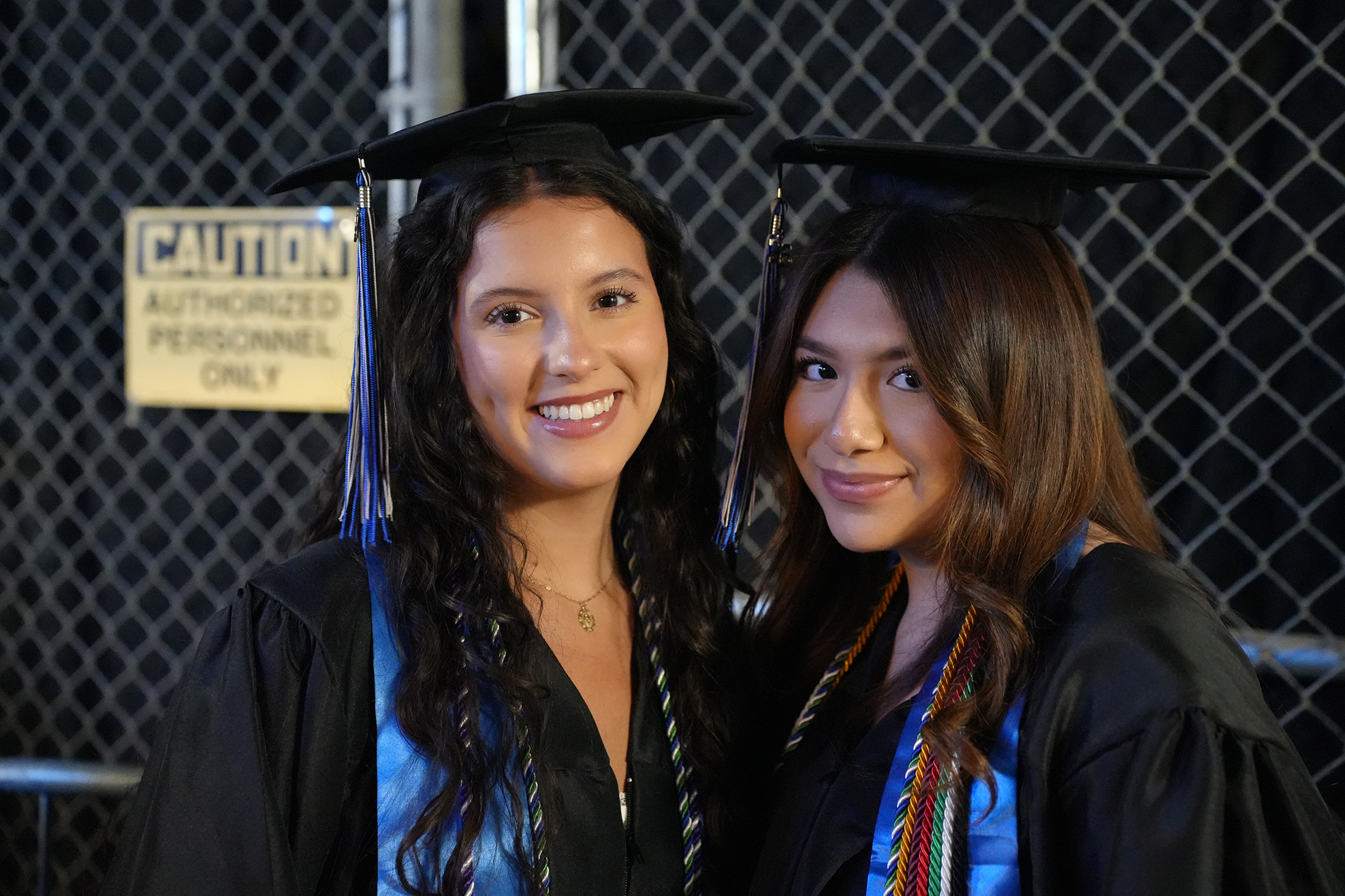 Two women in black gowns and hats stand close to each other in front of a metal fence.
