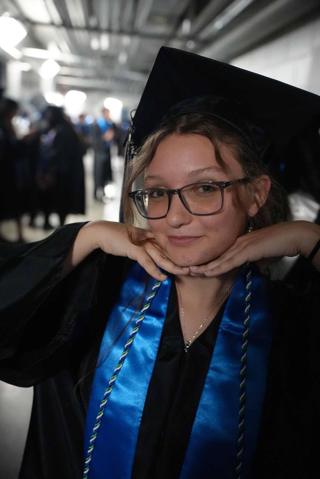 A woman in a graduation gown and cap, with glasses, smiling and posing with her hands on her chest.