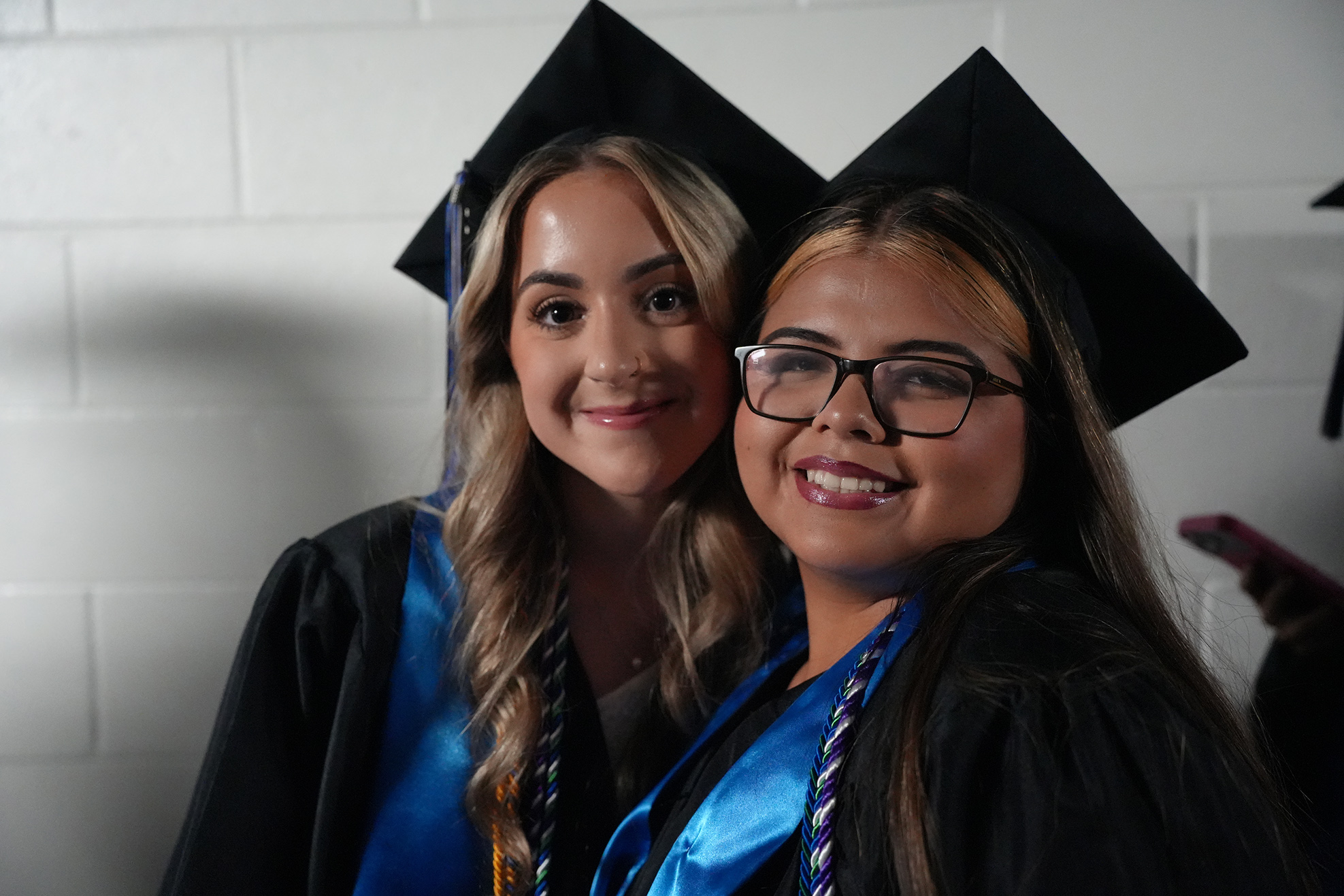 Two women in graduation robes and caps pose for a photo against a white brick wall.
