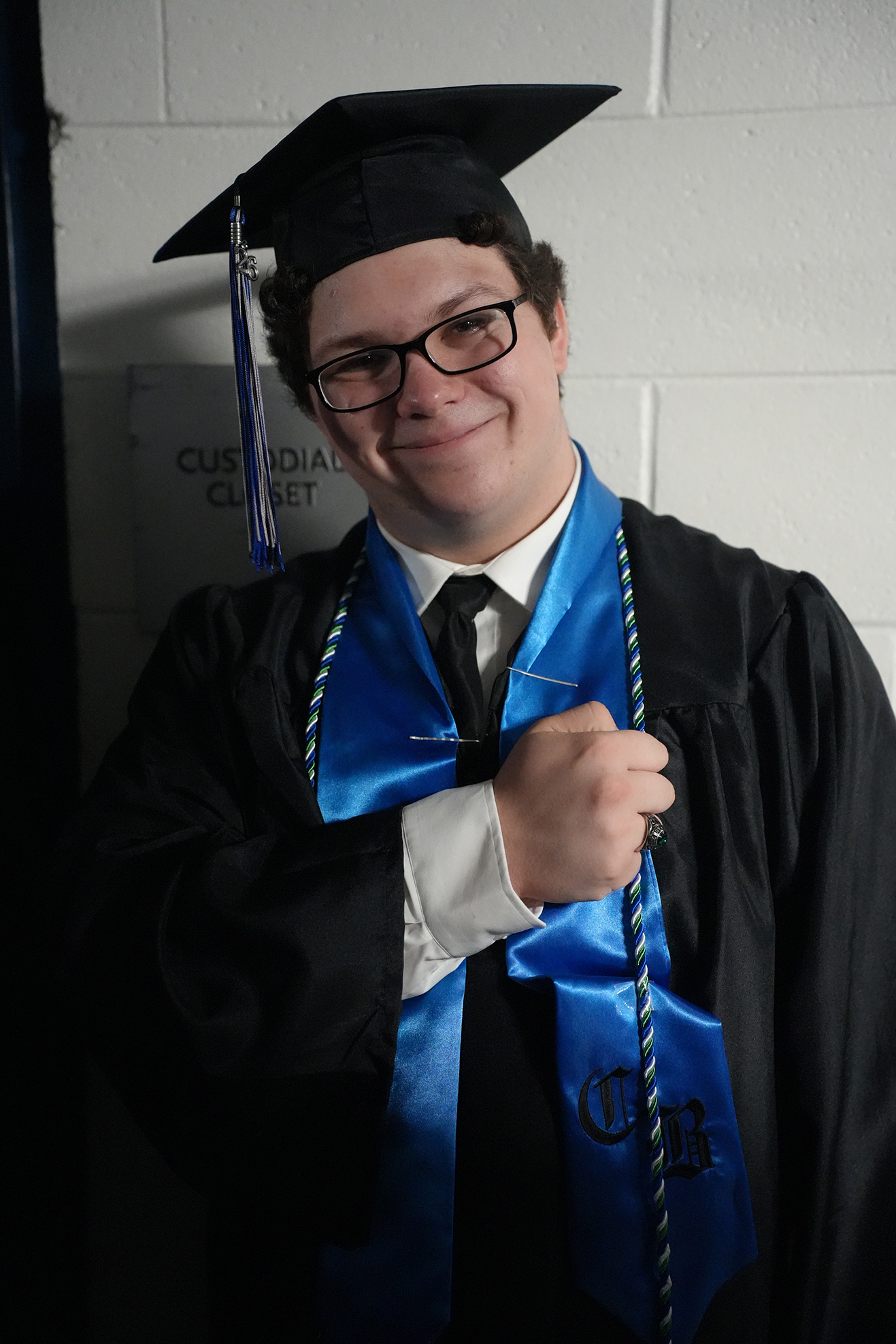A person in a graduation gown, hat, and glasses holds a tassel, smiling, against a white-walled background.