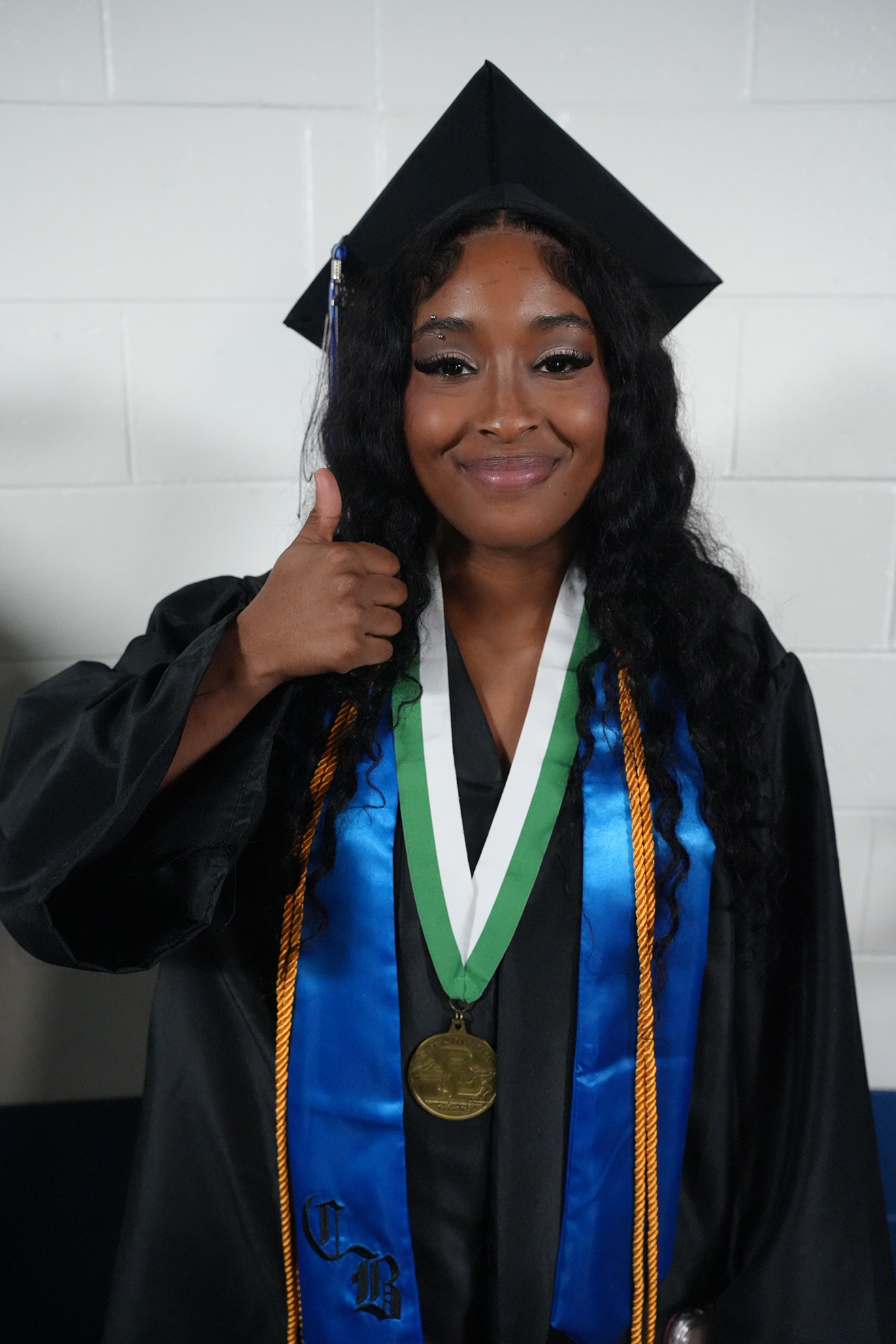 A woman in a black graduation gown and hat smiles while giving a thumbs-up gesture. She has a gold medal.