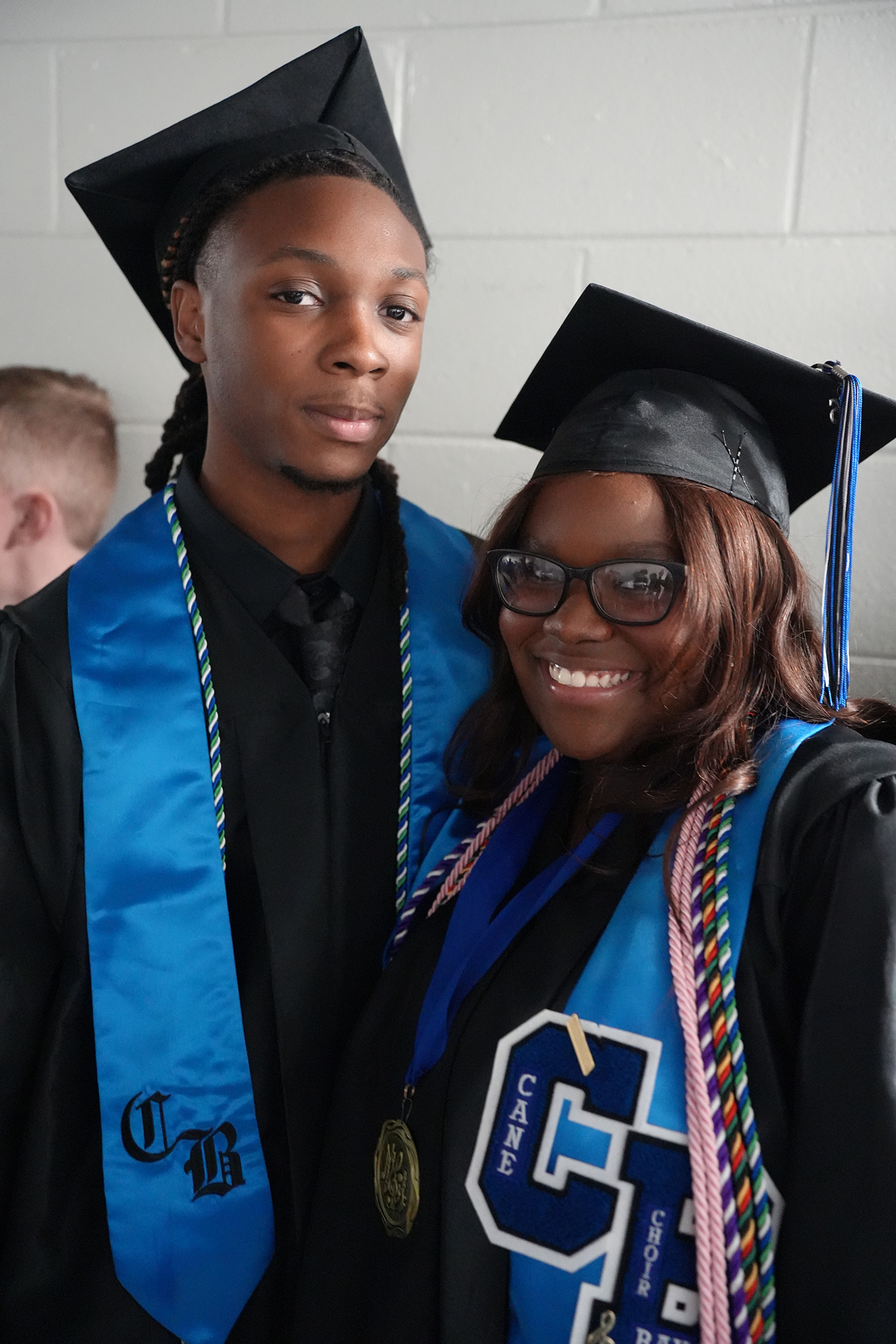 A young man and woman in black and blue graduation attire with blue stoles smile for the camera.
