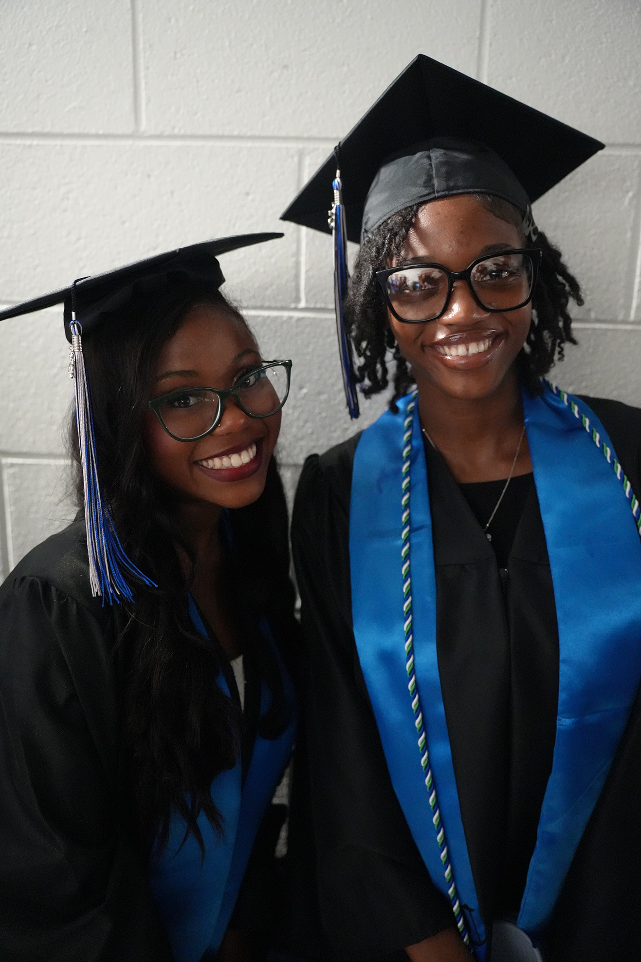 Two women in black graduation gowns and blue stoles. Both are smiling and wearing black caps with blue tassels.