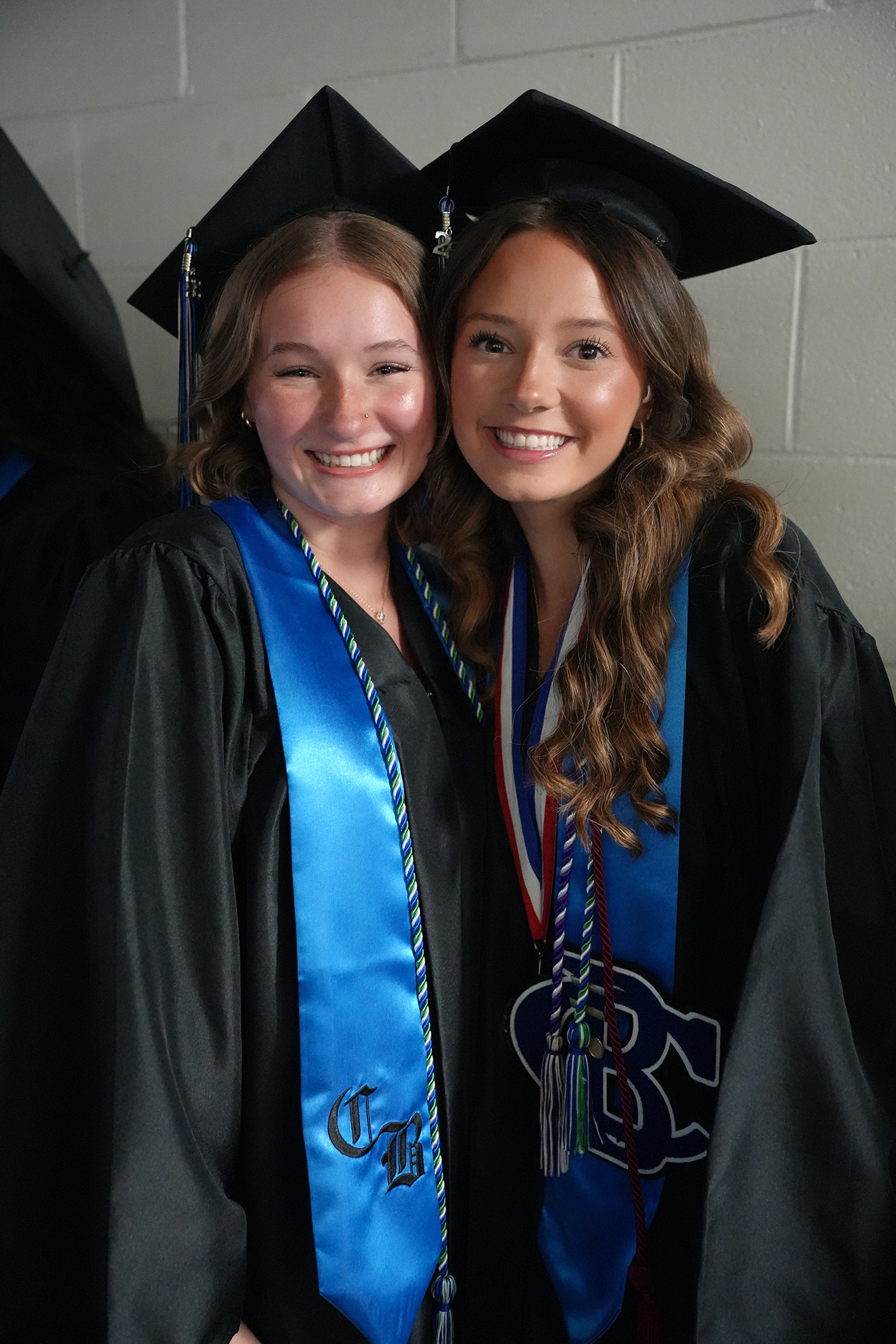 Two women in black gowns and blue stoles, one with a sash reading "TH," smiling against a light-colored wall.