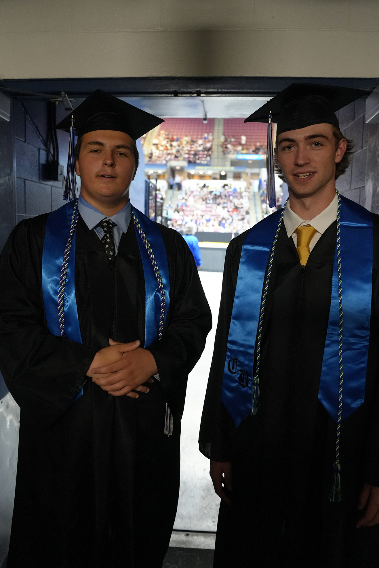 Two men in academic regalia stand in a corridor, with a blurred stadium visible in the background.