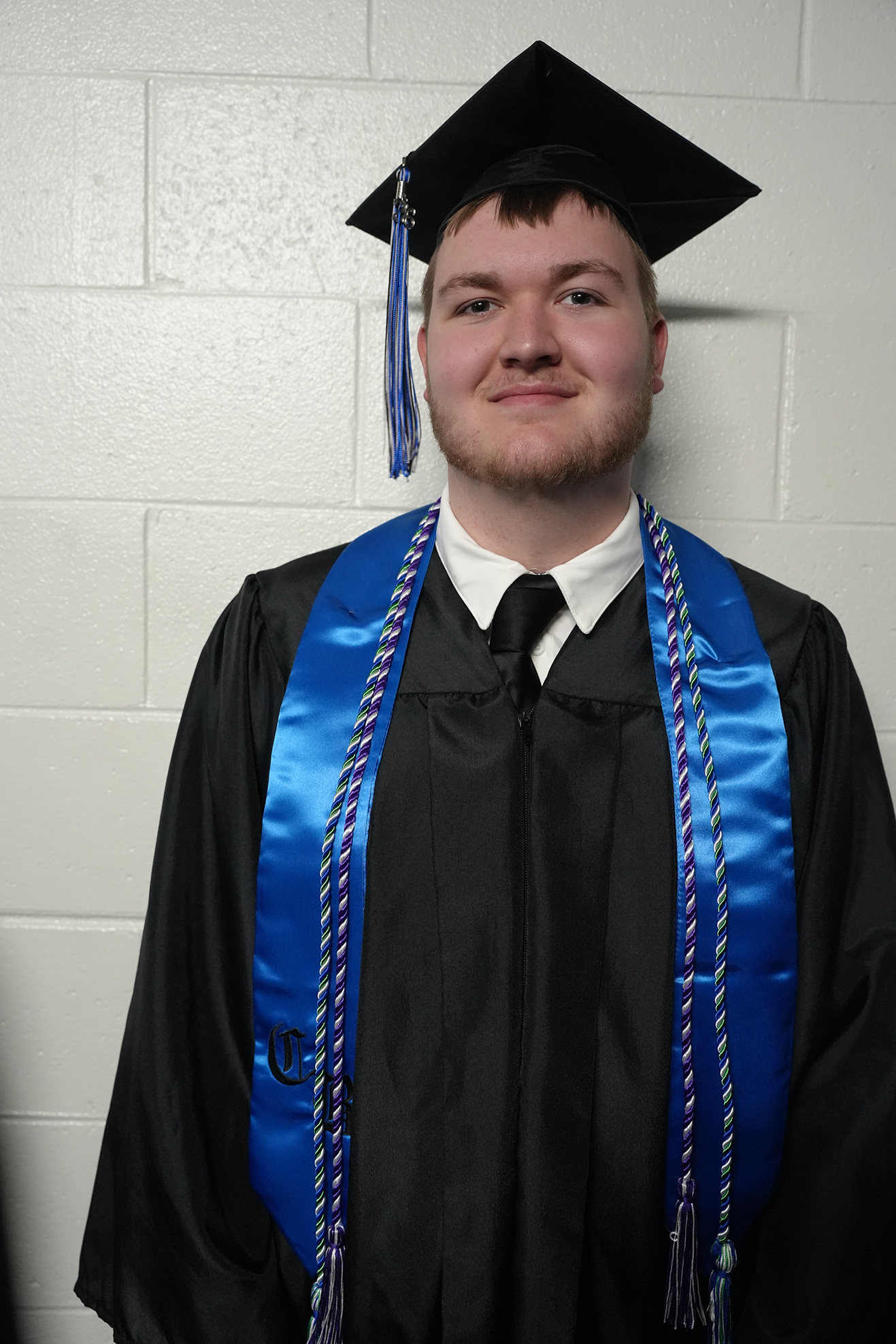 A man in a black graduation gown with a blue stole stands against a white brick wall, wearing a cap with blue tassels.