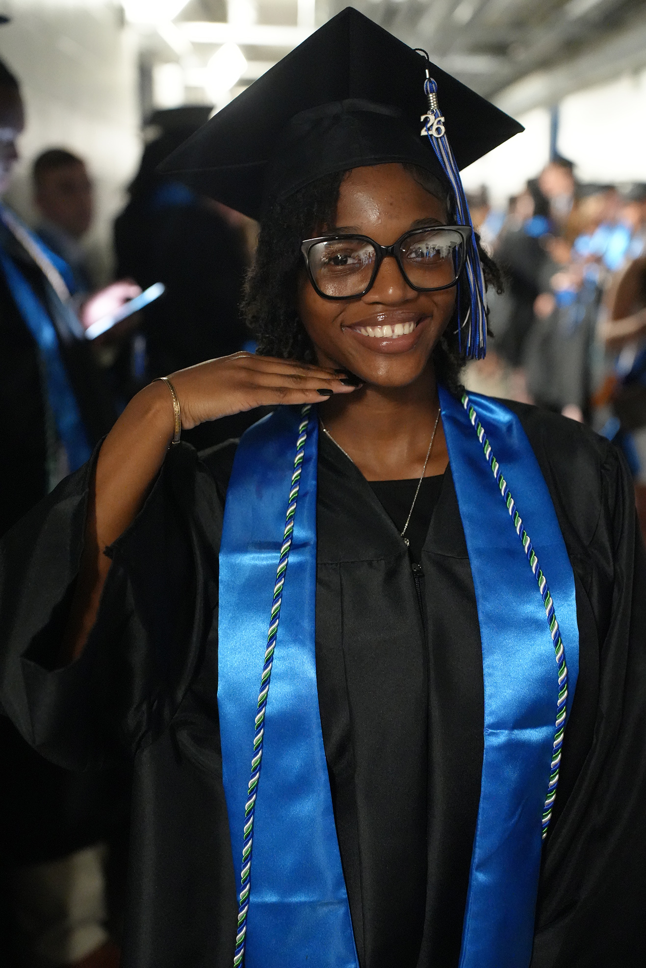 A smiling woman in a black graduation gown and blue stole with a tassel. She wears glasses and a graduation cap.