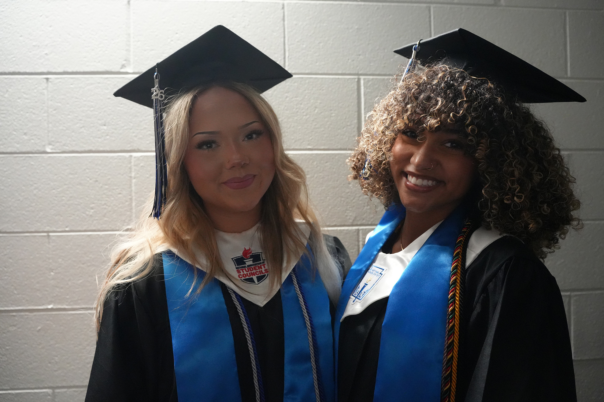 Two women wearing graduation gowns and caps stand together, smiling. One has blonde hair; the other has curly hair.