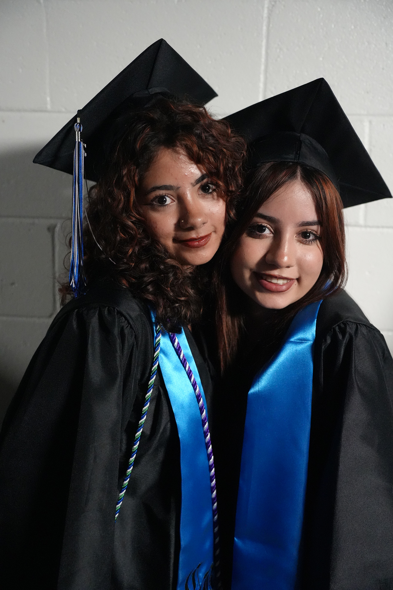 Two women in black graduation gowns and hats, standing close together. One has a blue sash, the other has a multicolored sash.