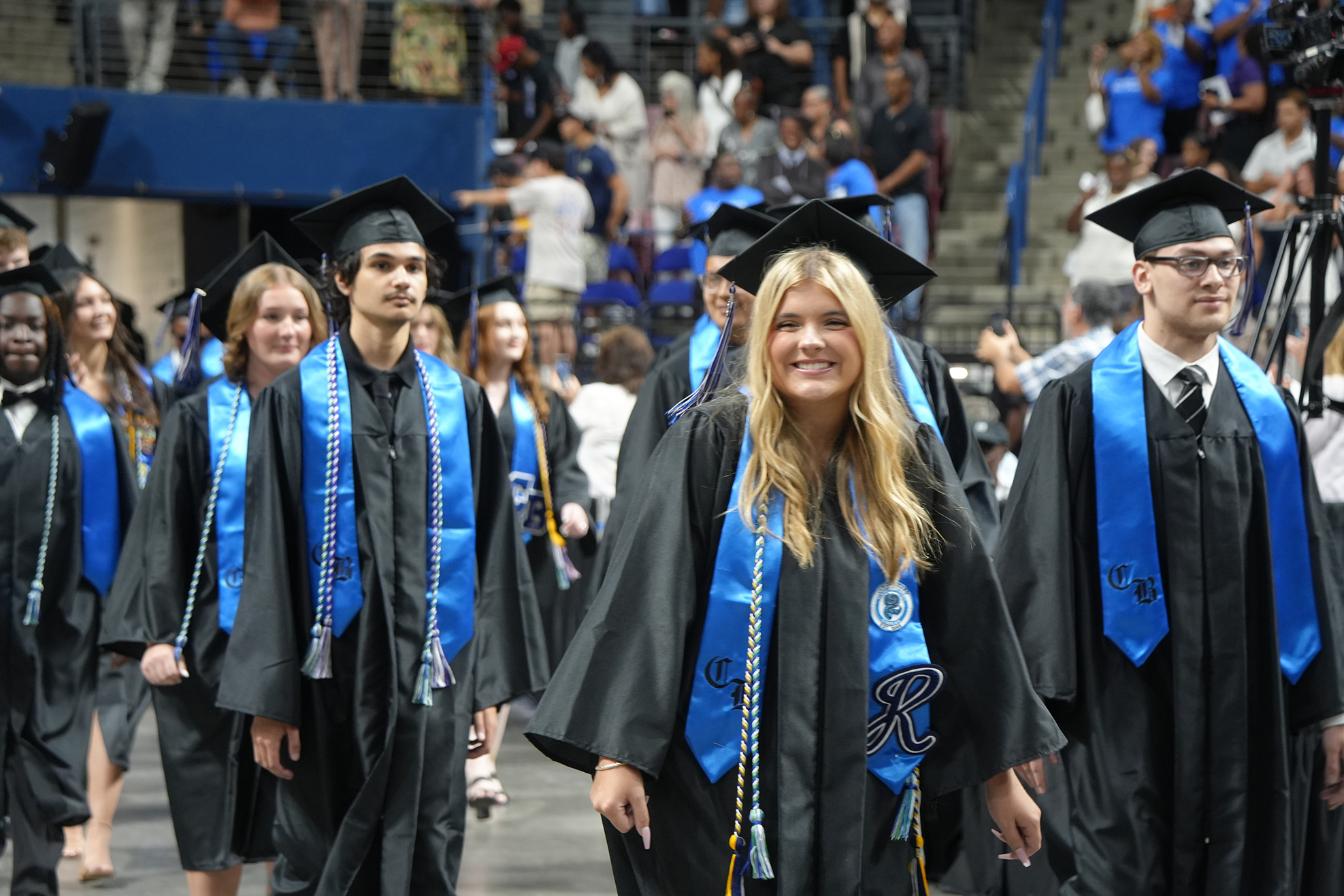 Graduates in black gowns and blue stoles walk down a stadium aisle with an audience seated in the background.