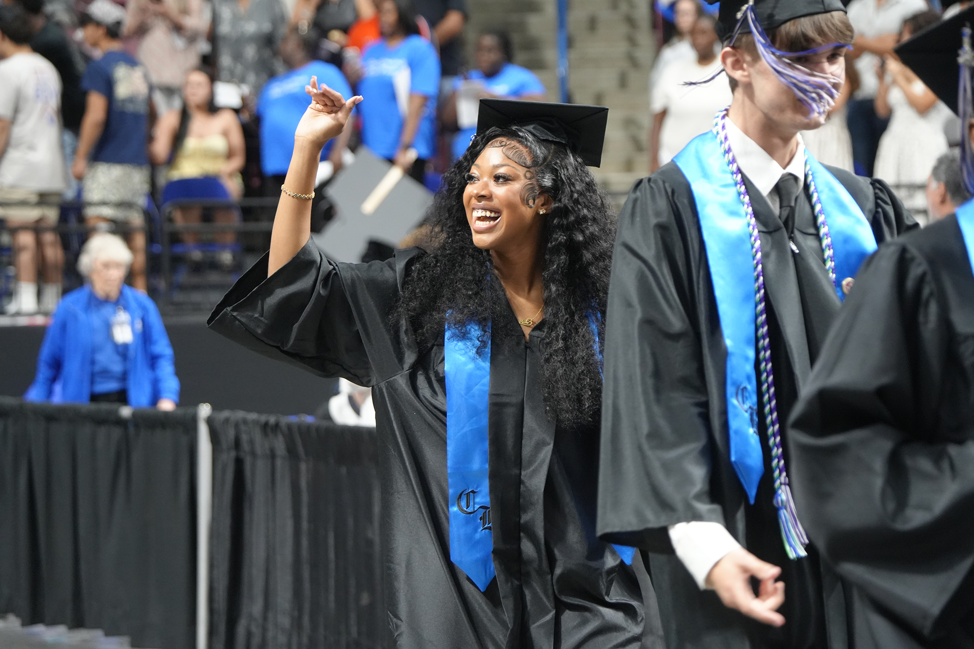 A smiling woman in a cap and gown celebrates while walking with two others in front of a crowd.