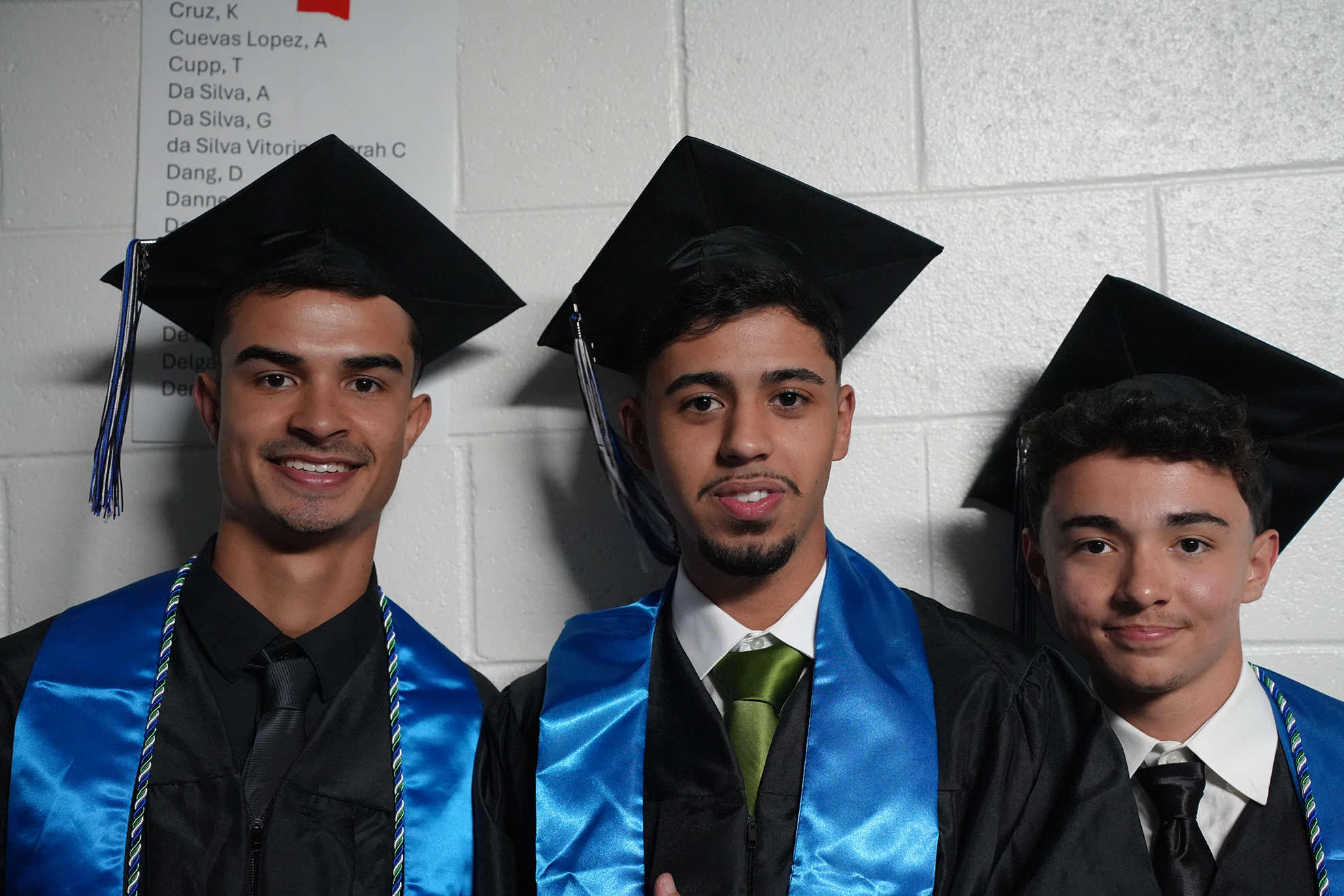 Three individuals in black graduation gowns and blue stoles, with one wearing a green tie, stand close together, smiling.