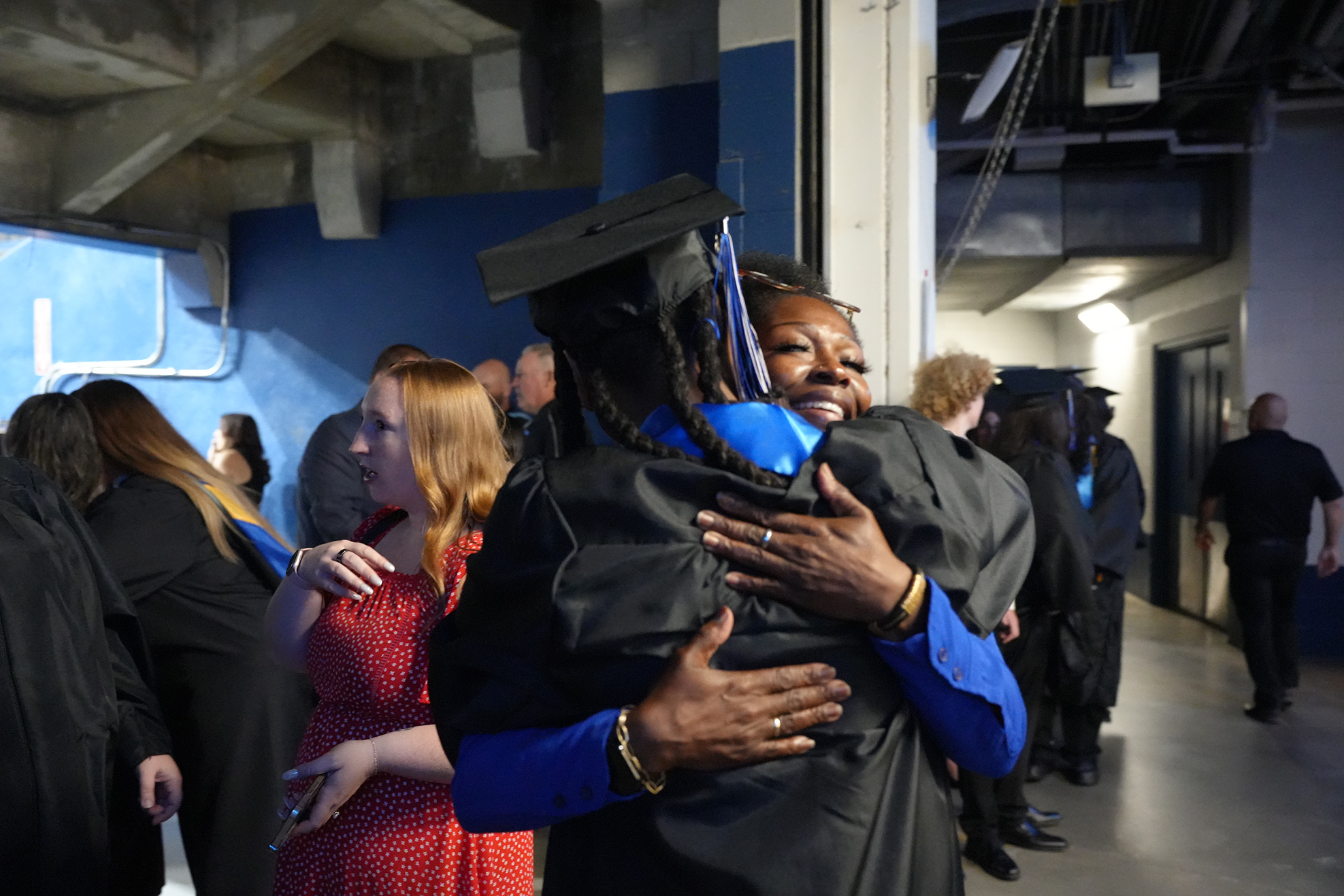 People gather in an indoor area, possibly a school. A woman hugs another in a cap and gown.