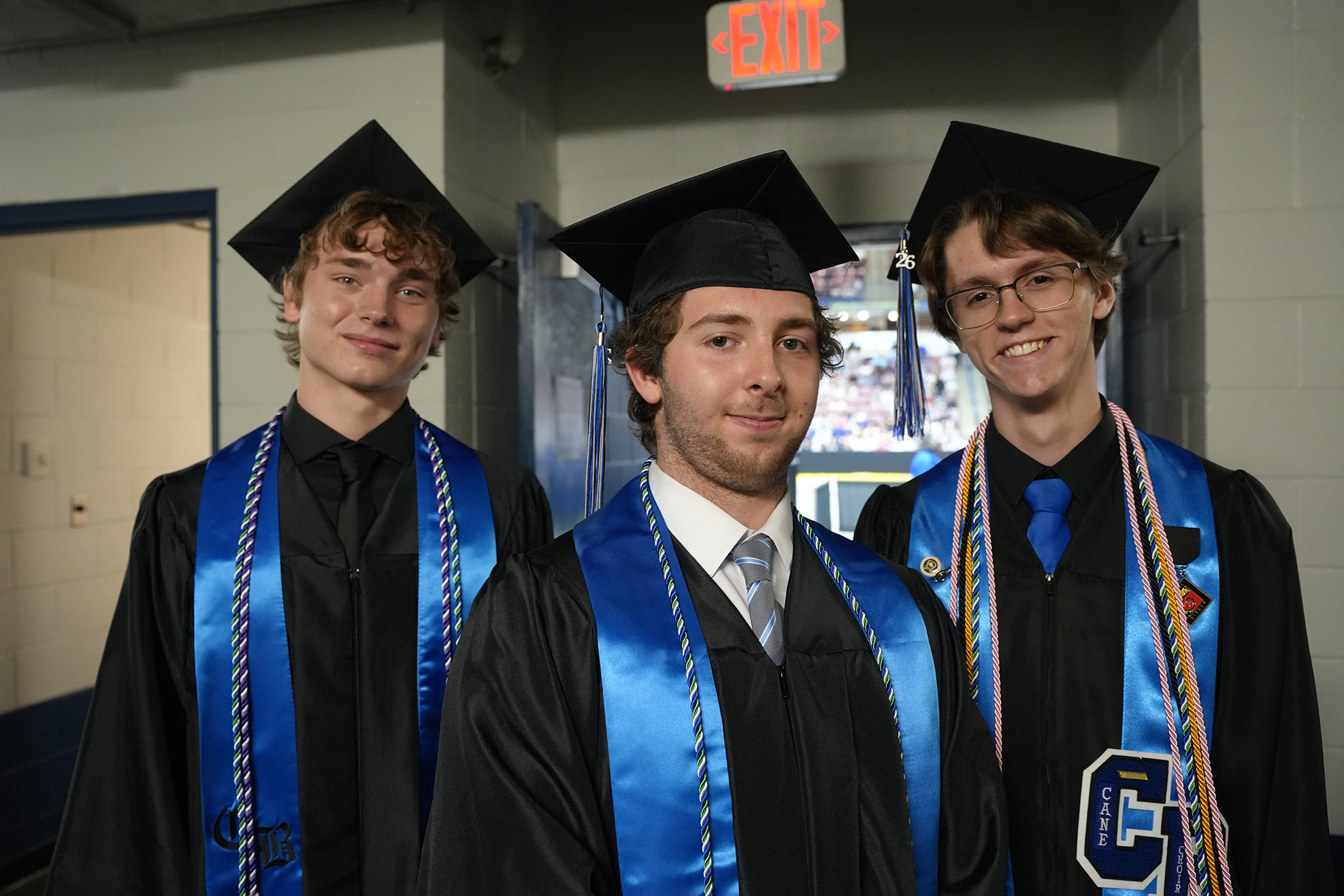 Three men in graduation gowns, hats, and ties stand in a corridor. One wears glasses.