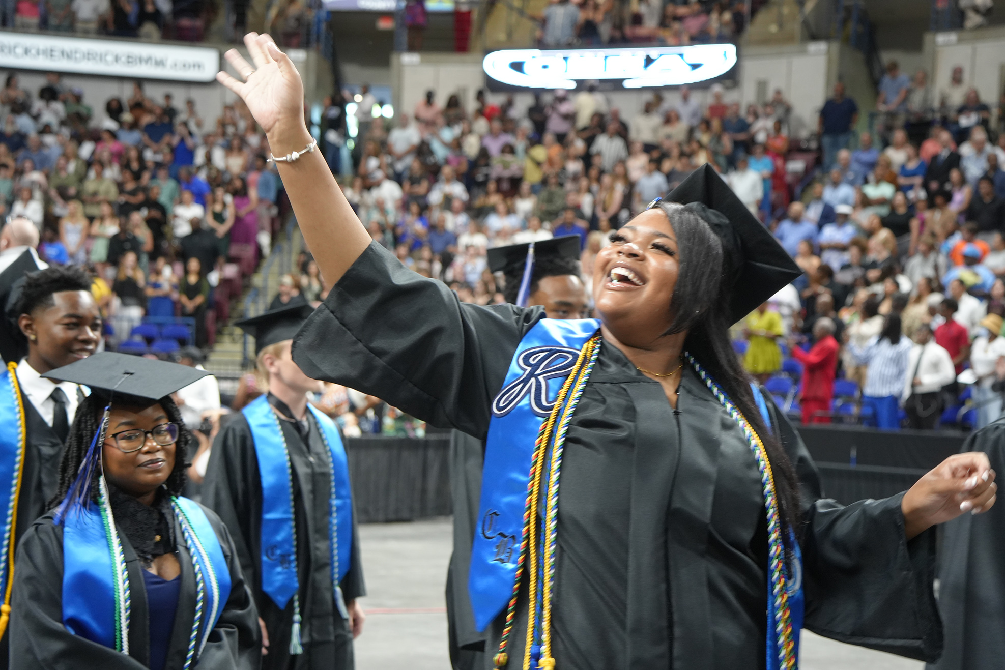 A woman in a black gown and cap waves while walking with others at a graduation ceremony.