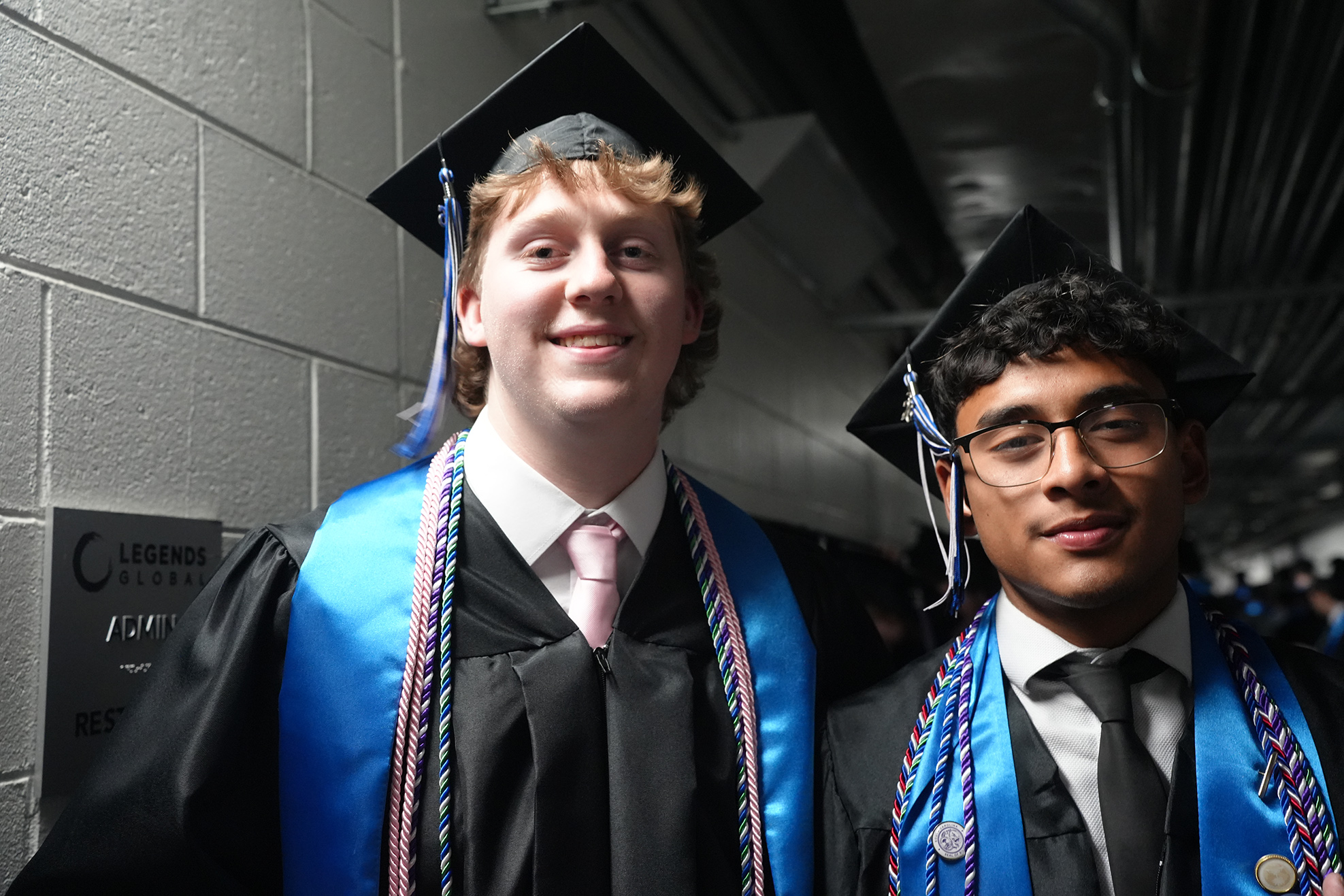 Two young men in graduation robes and caps stand close to a wall with a sign.