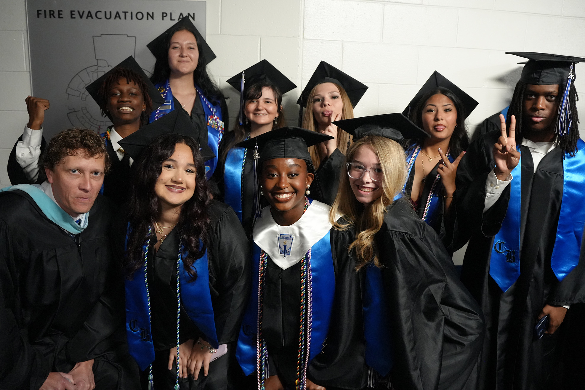 A group of people in black graduation gowns with blue sashes pose for a photo in front of a white background.