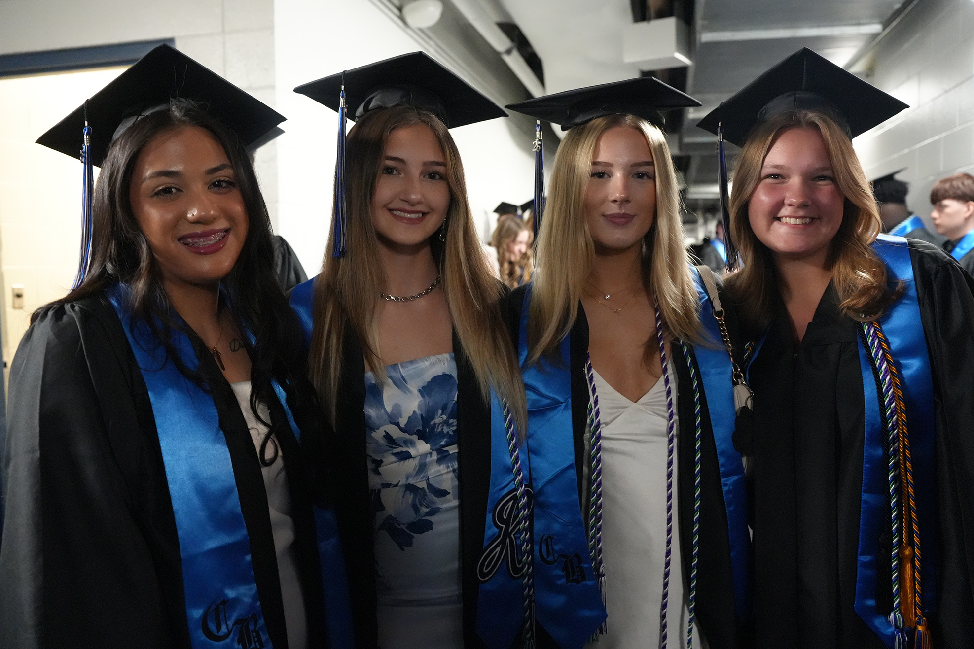 Four women in graduation gowns and caps, smiling. One wears a blue floral dress. Two others wear sleeveless white and blue dresses.