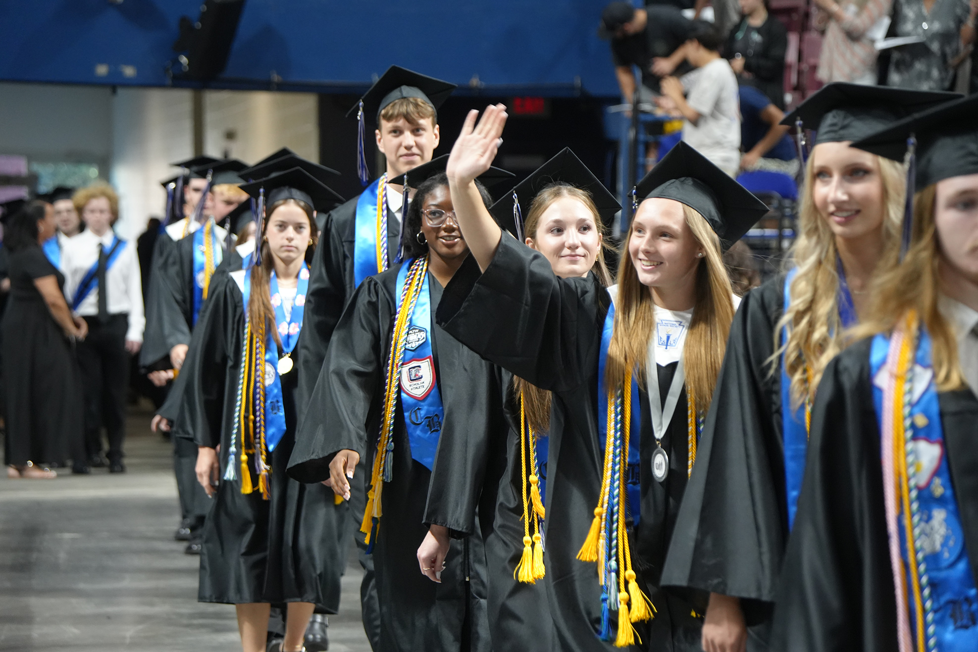Graduates in black gowns and caps walk in a line. They wave at the audience.