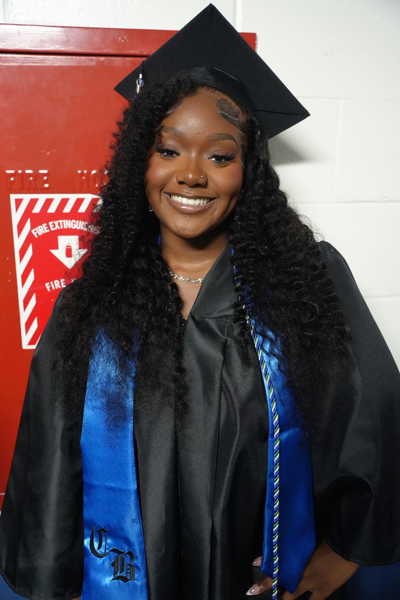 Person with curly hair, wearing a graduation gown, cap, and sash. Standing in front of a fire extinguisher locker.