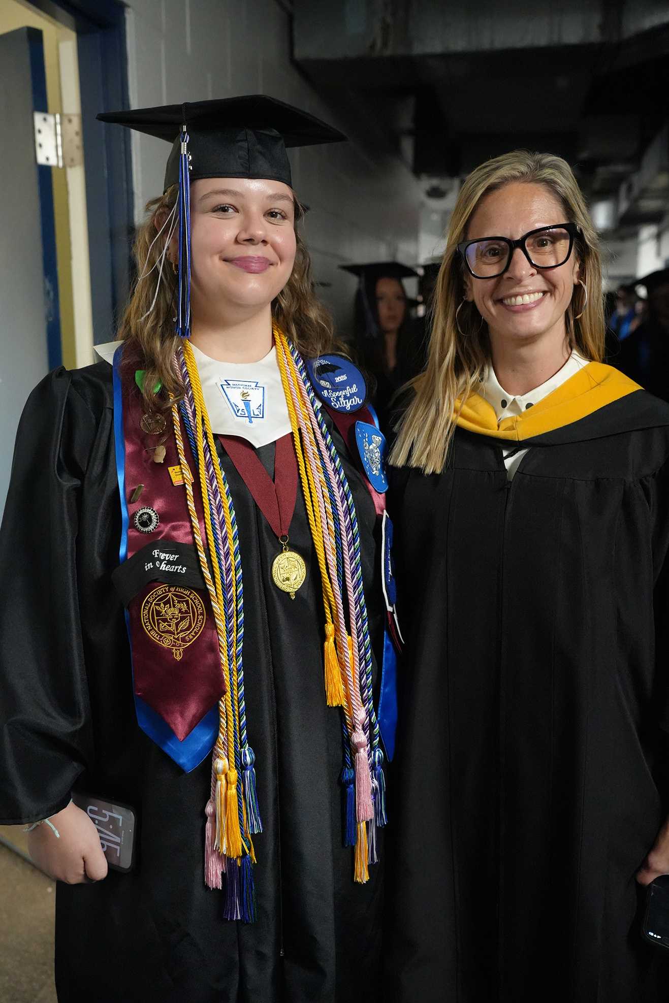 Two women in graduation gowns, one with a cap and a sash adorned with multicolored cords and medals, pose for a photo.