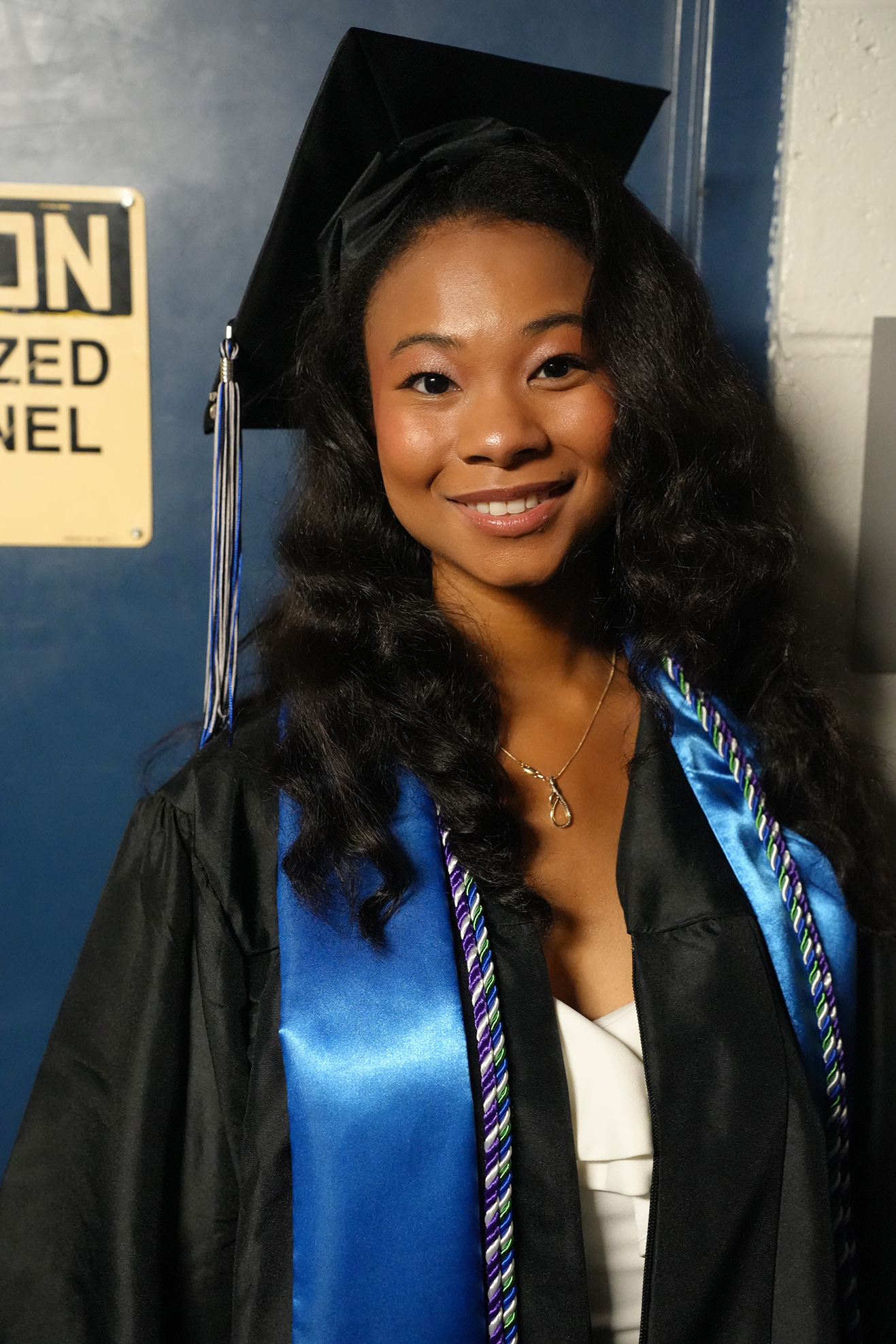 A person with curly hair in a gown, smiling. The gown is black with blue and multicolored sashes.