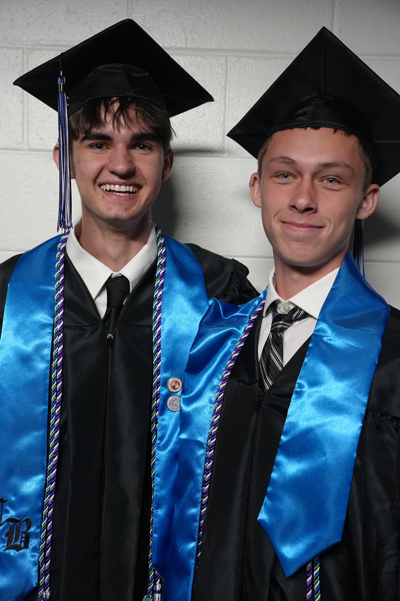 Two men in caps and gowns standing together against a white brick wall. Both wear blue stoles with purple and white tassels.