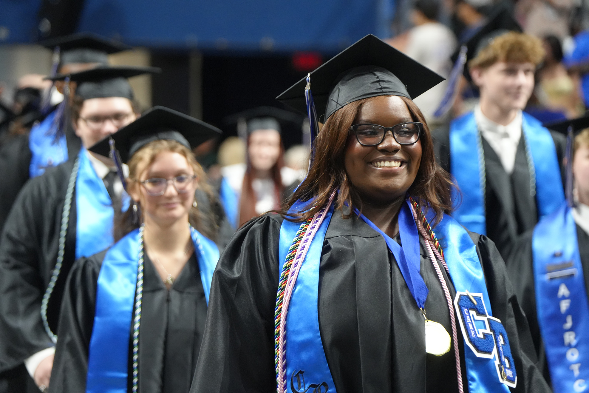 A woman wearing a black graduation gown with a blue stole smiles at the camera during a graduation ceremony.