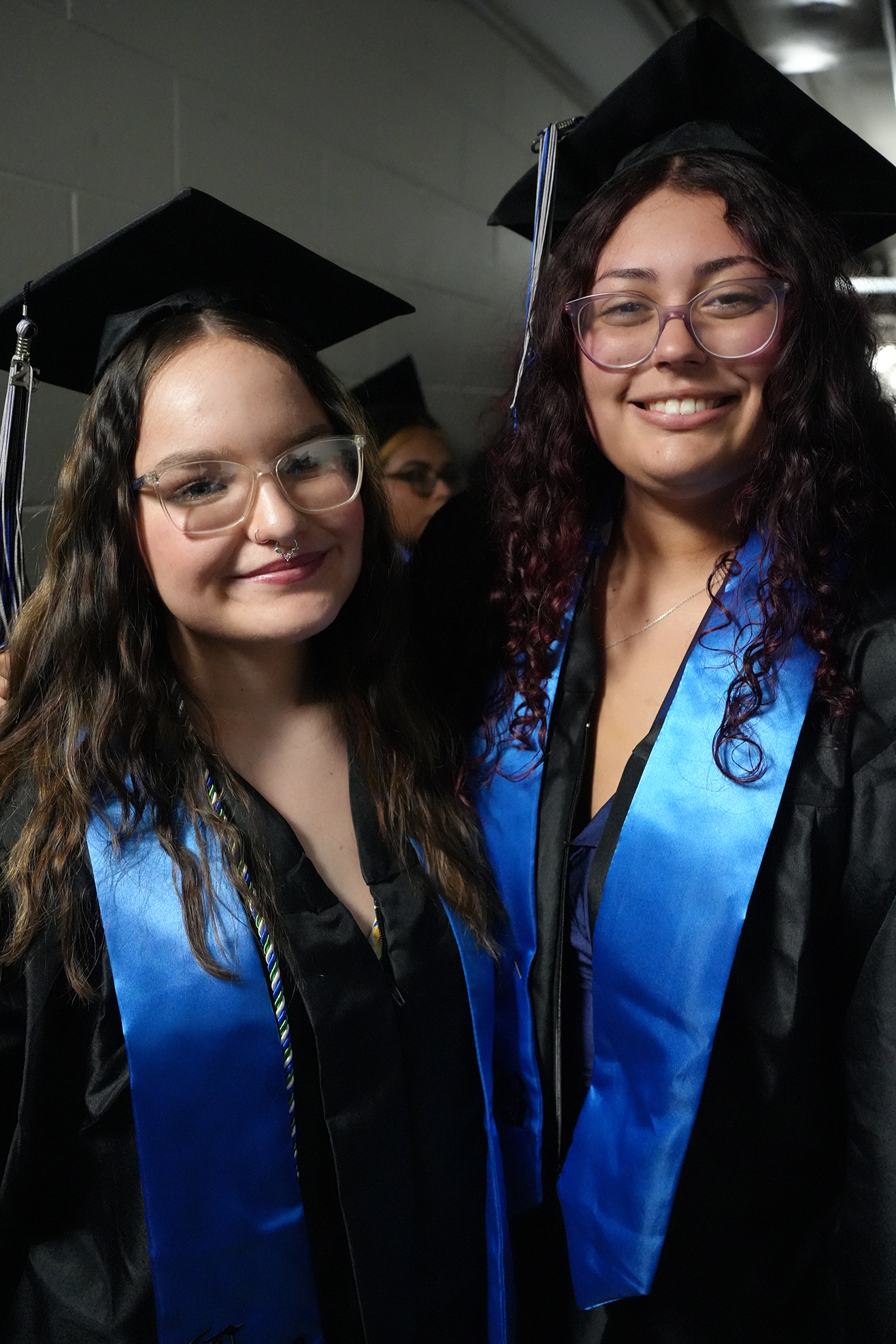 Two women in graduation robes and hats stand in a corridor. Both wear glasses and smile. Behind them, a blurred figure is visible.
