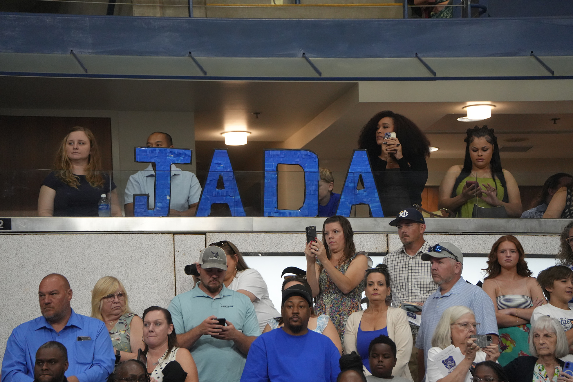 Spectators in a sports venue, many using phones, stand behind a banner reading "JADA".
