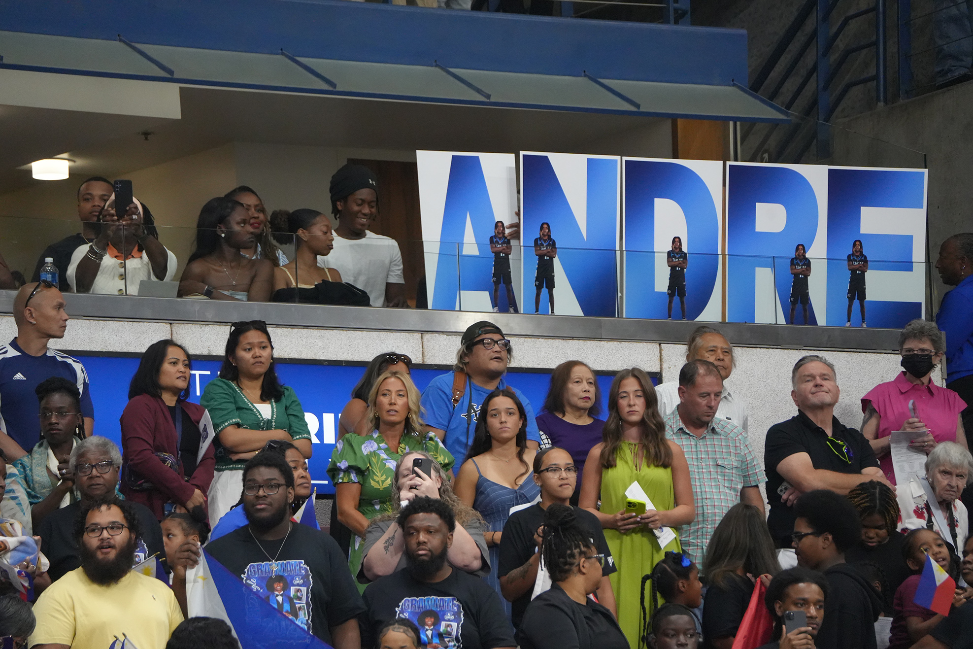Audience in a stadium, watching an event. Some hold phones to record, and a banner says "ANDRE" above.