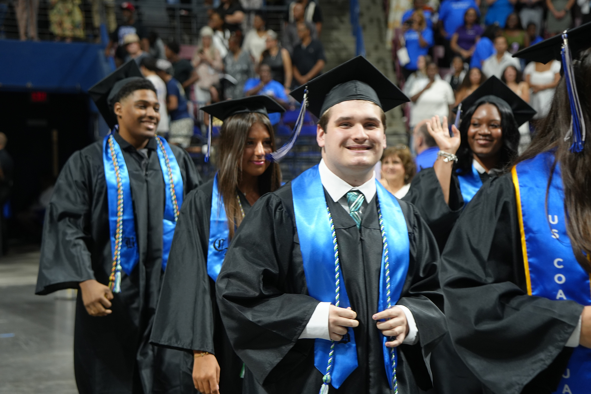 Graduates in blue and black gowns and caps walk across the stage at an event.